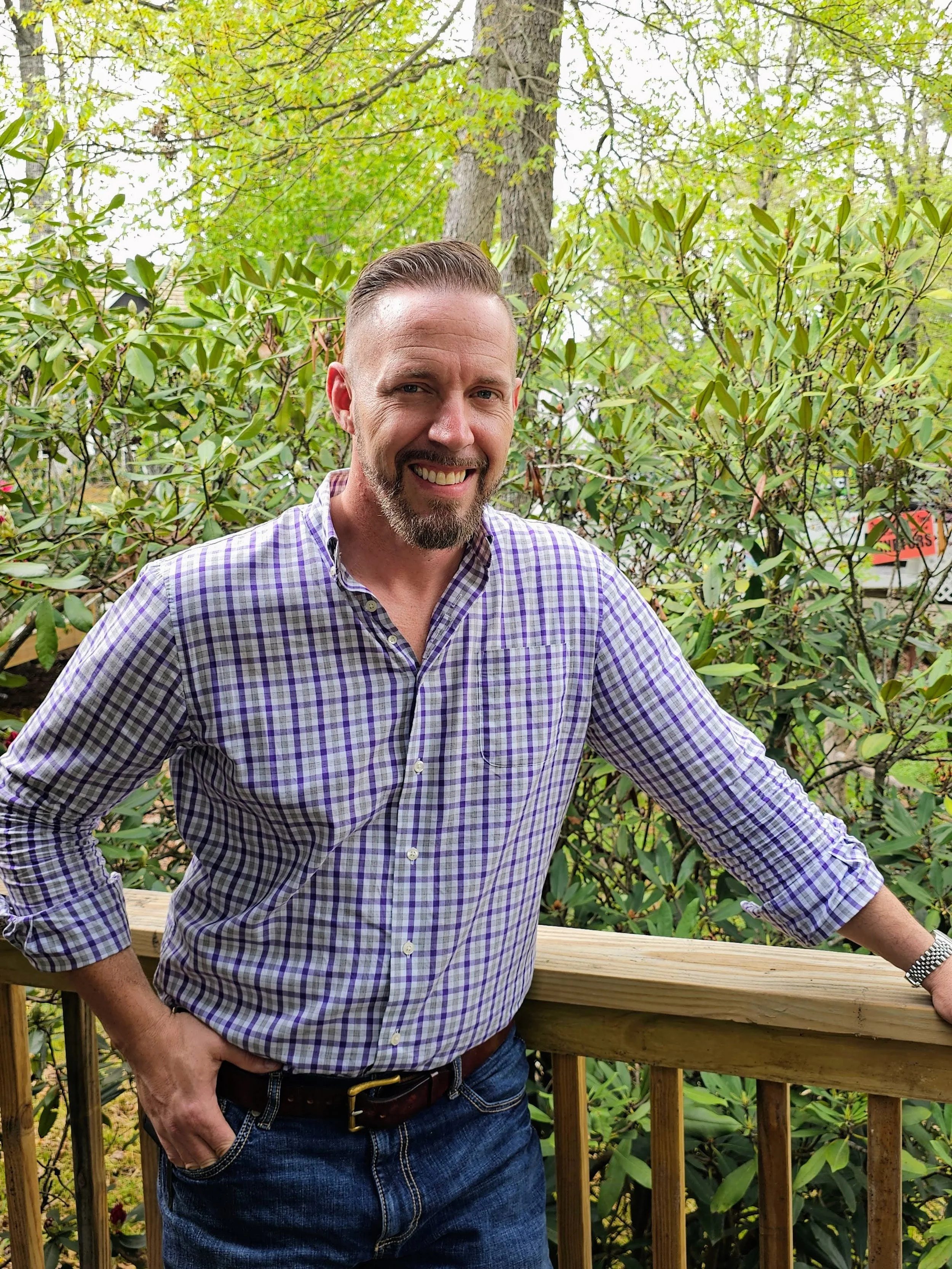 A smiling man with a beard and mustache wearing a purple checked shirt and jeans, standing outdoors on a wooden deck with a background of green foliage, trees, and plants.