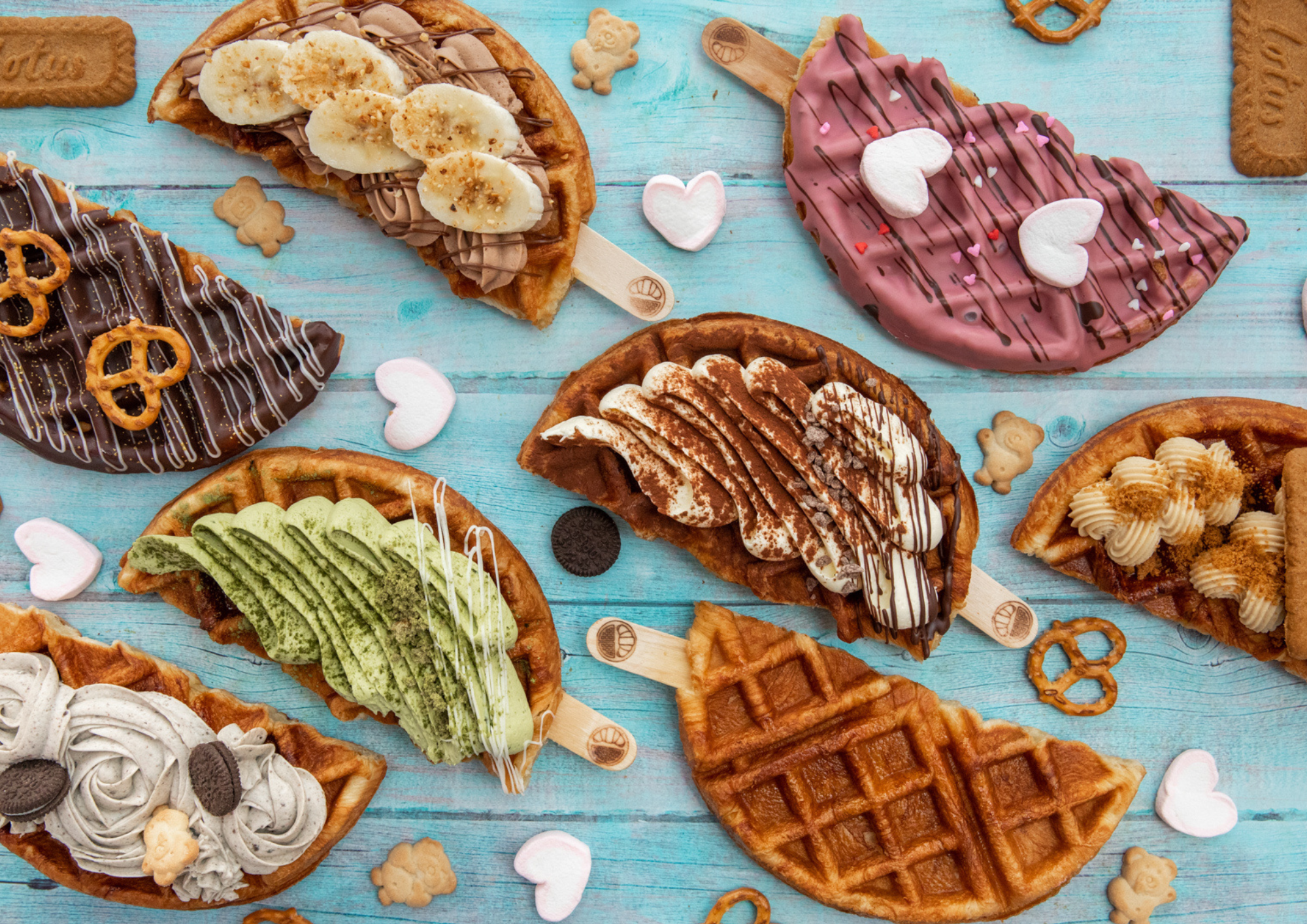 Variety of decorated waffle ice cream tacos with different toppings and flavors on a blue wooden surface, decorated with small heart and teddy bear-shaped treats.