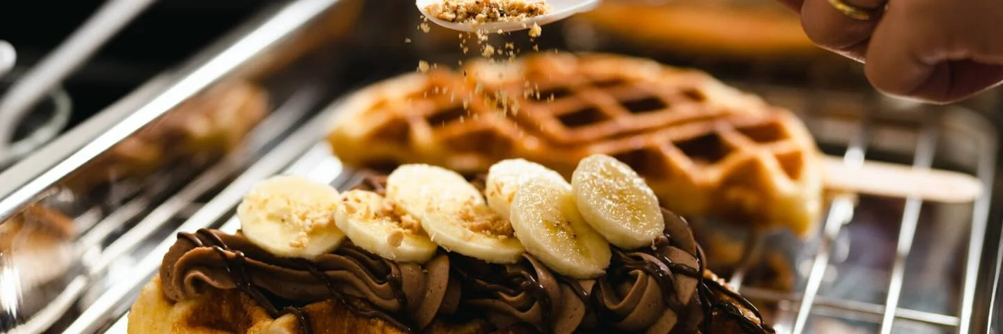 A waffle topped with sliced bananas and chocolate frosting, with a scene of a waffle iron in the background.