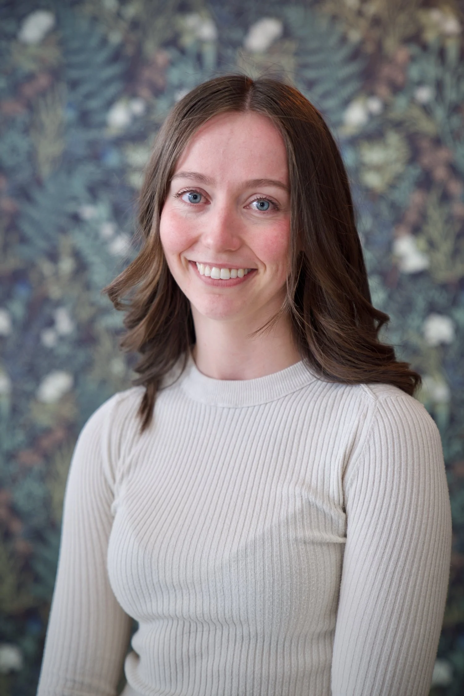 A young woman with shoulder-length brown hair, blue eyes, and a bright smile, wearing a cream-colored ribbed long-sleeve top, standing in front of a blurred floral background.