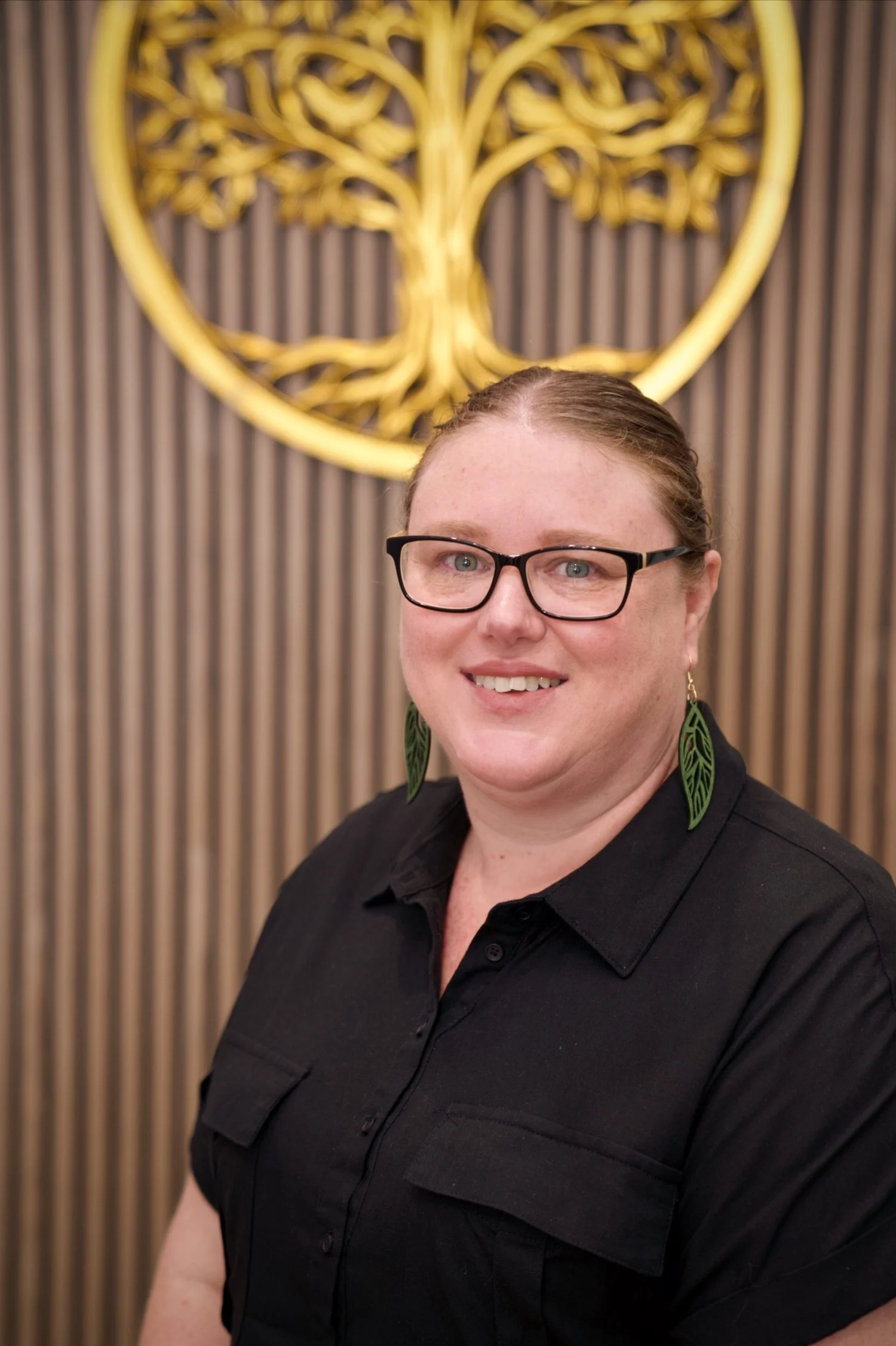 A woman wearing glasses and green leaf-shaped earrings, smiling, standing in front of a wooden wall with a gold tree emblem.