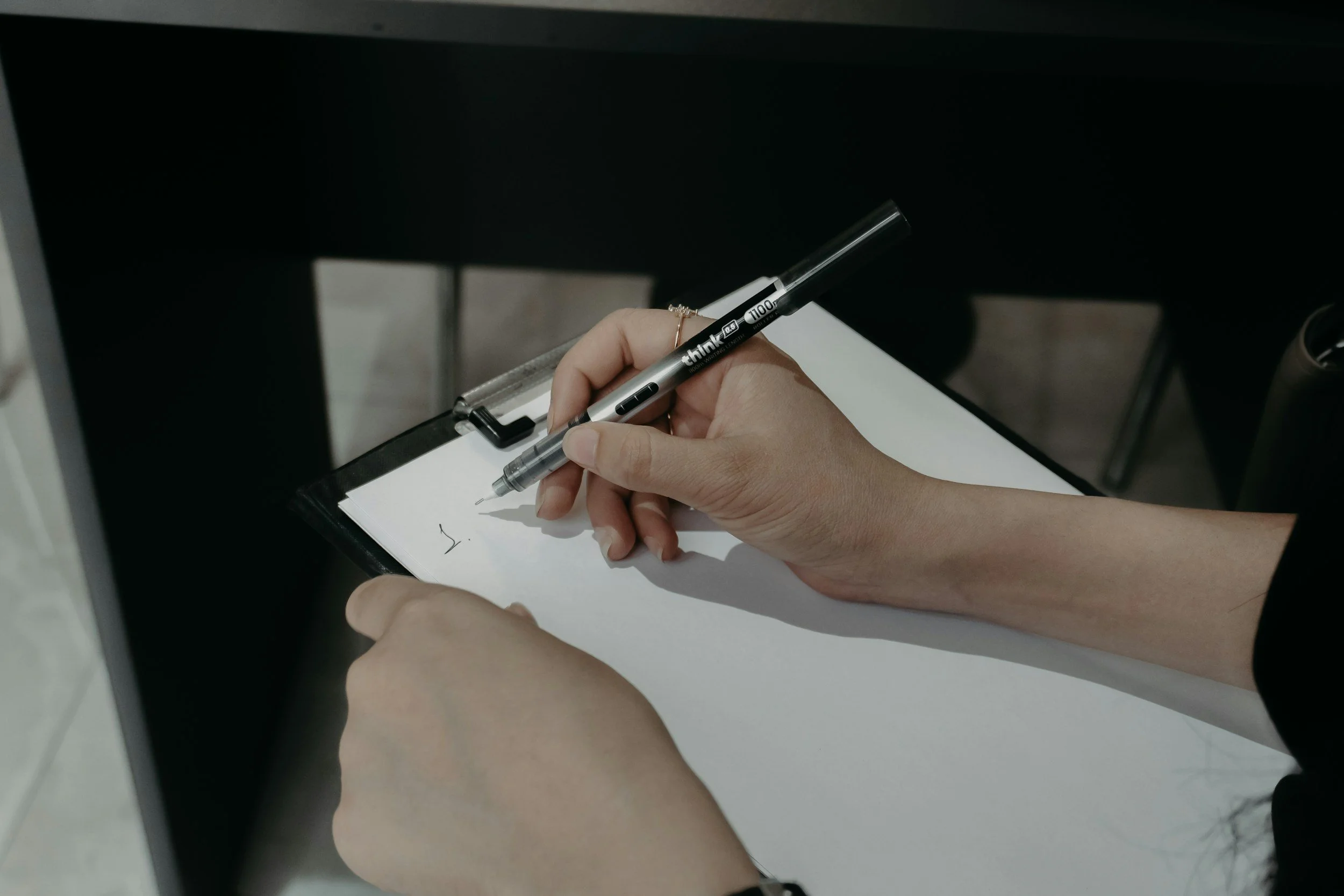 A person writing on a notepad with a black ballpoint pen at a desk.