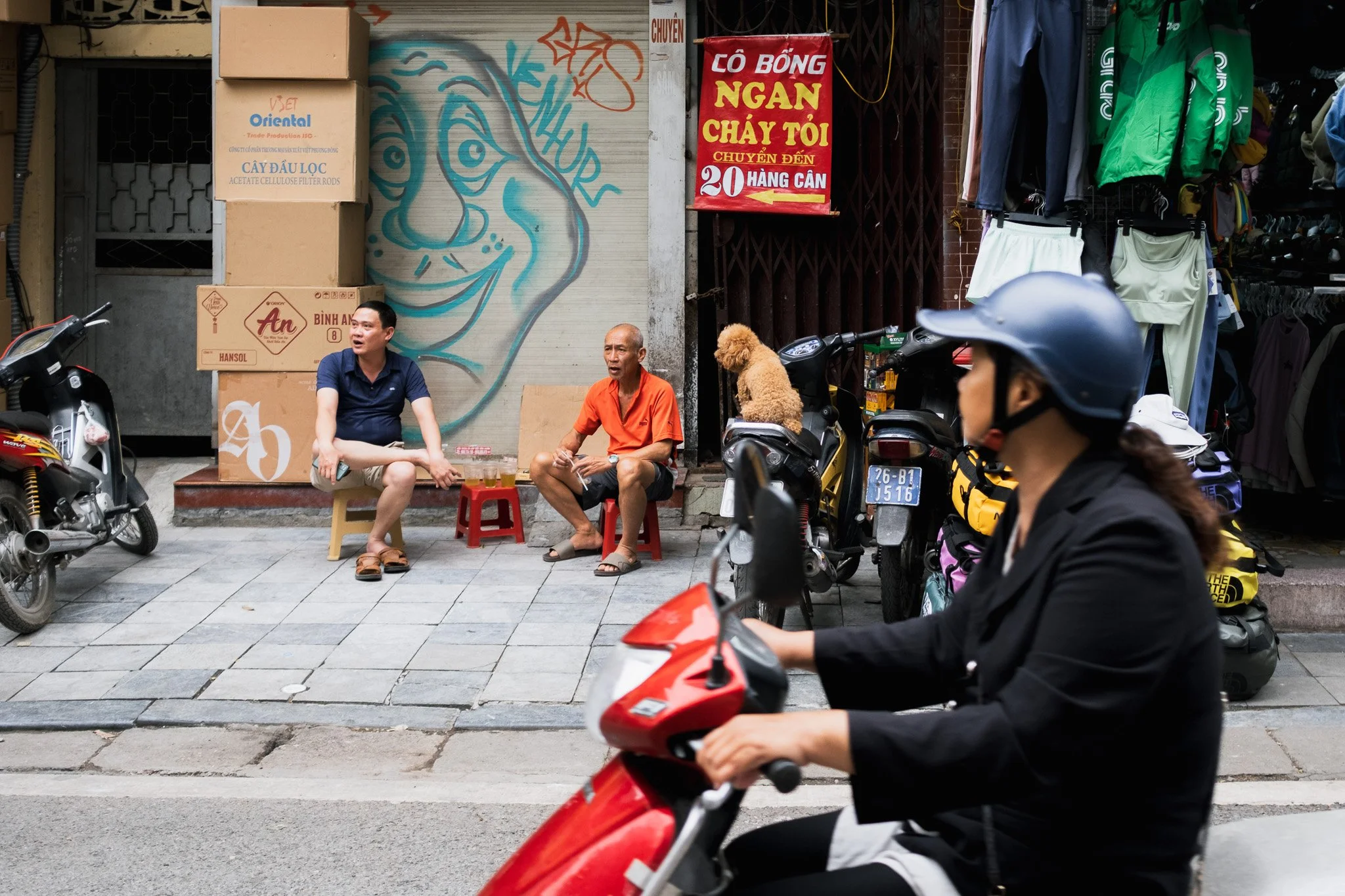 Two men sitting on small stools on a sidewalk, having drinks, with a mural of a face in the background. A woman on a scooter passes by, and there are parked motorcycles and a clothing stall nearby.
