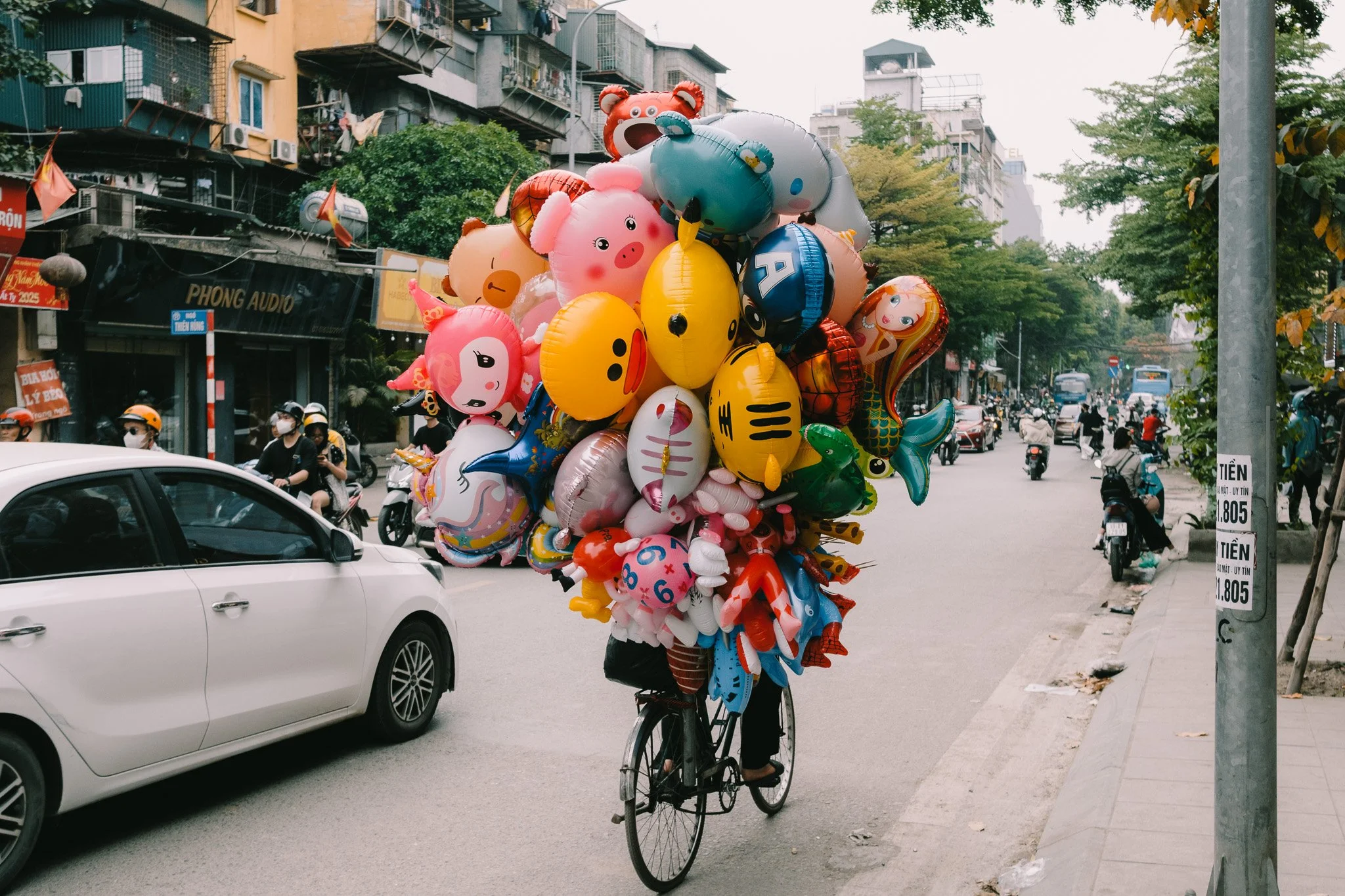 A street scene with a person riding a bicycle loaded with colorful balloons shaped like animals and objects.