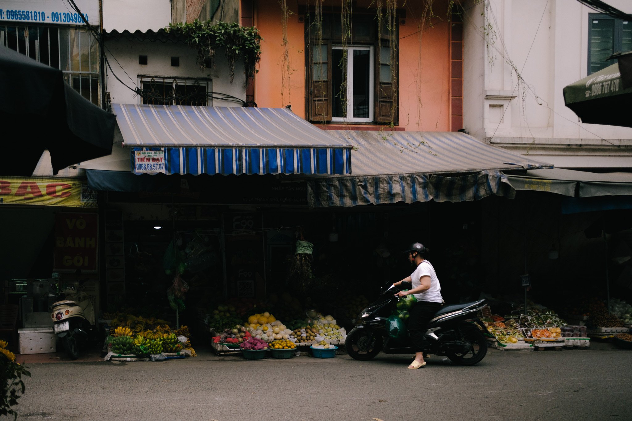 A woman on a black scooter in front of a fruit stand with various fruits on display, in an urban street scene under awnings.