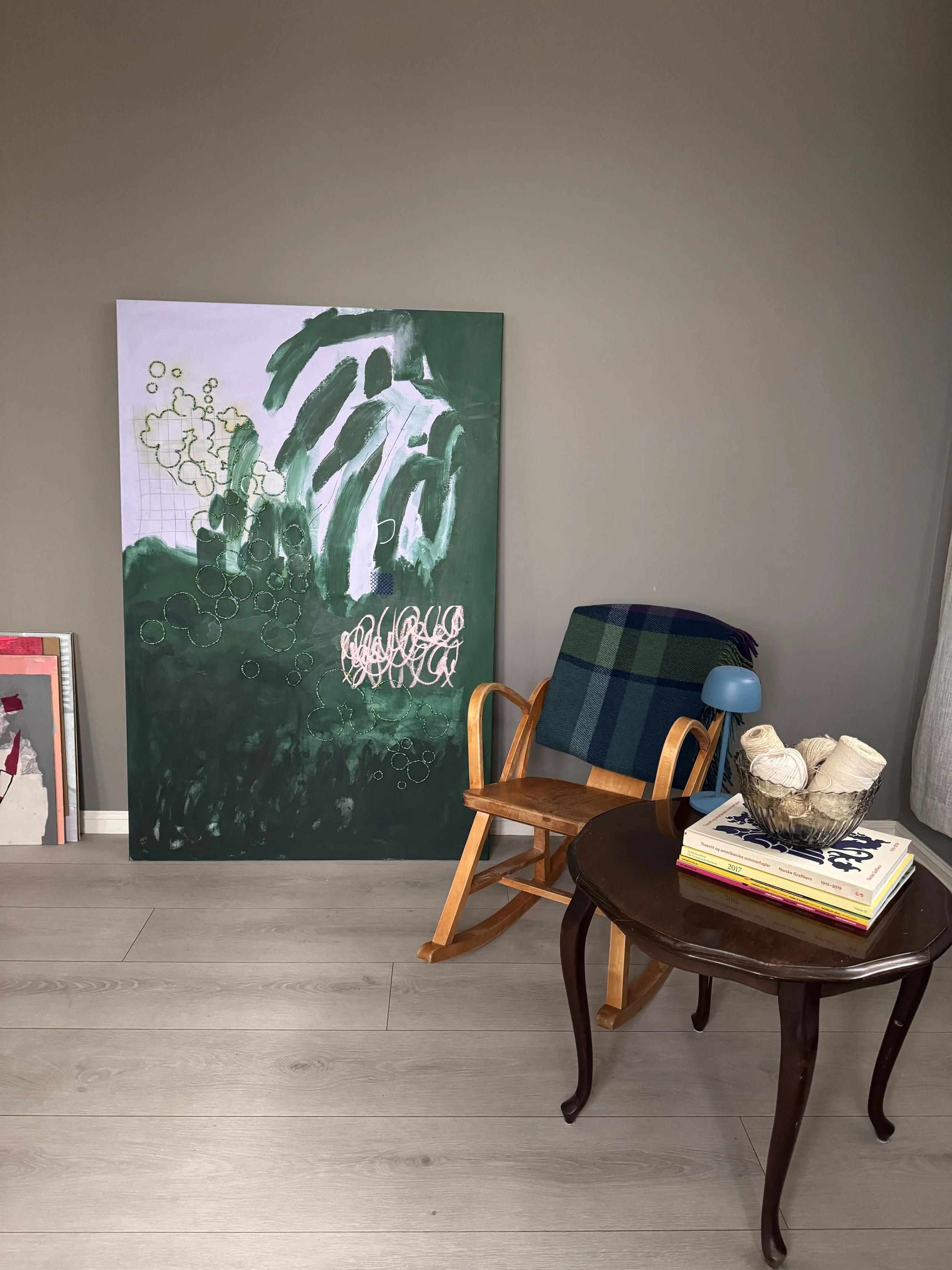 Living room corner with a dark wooden round table, a stack of books, and a bowl of yarn. A wooden chair and a plaid upholstered chair with a blue lamp are nearby, with a large green and white abstract painting on the beige wall.