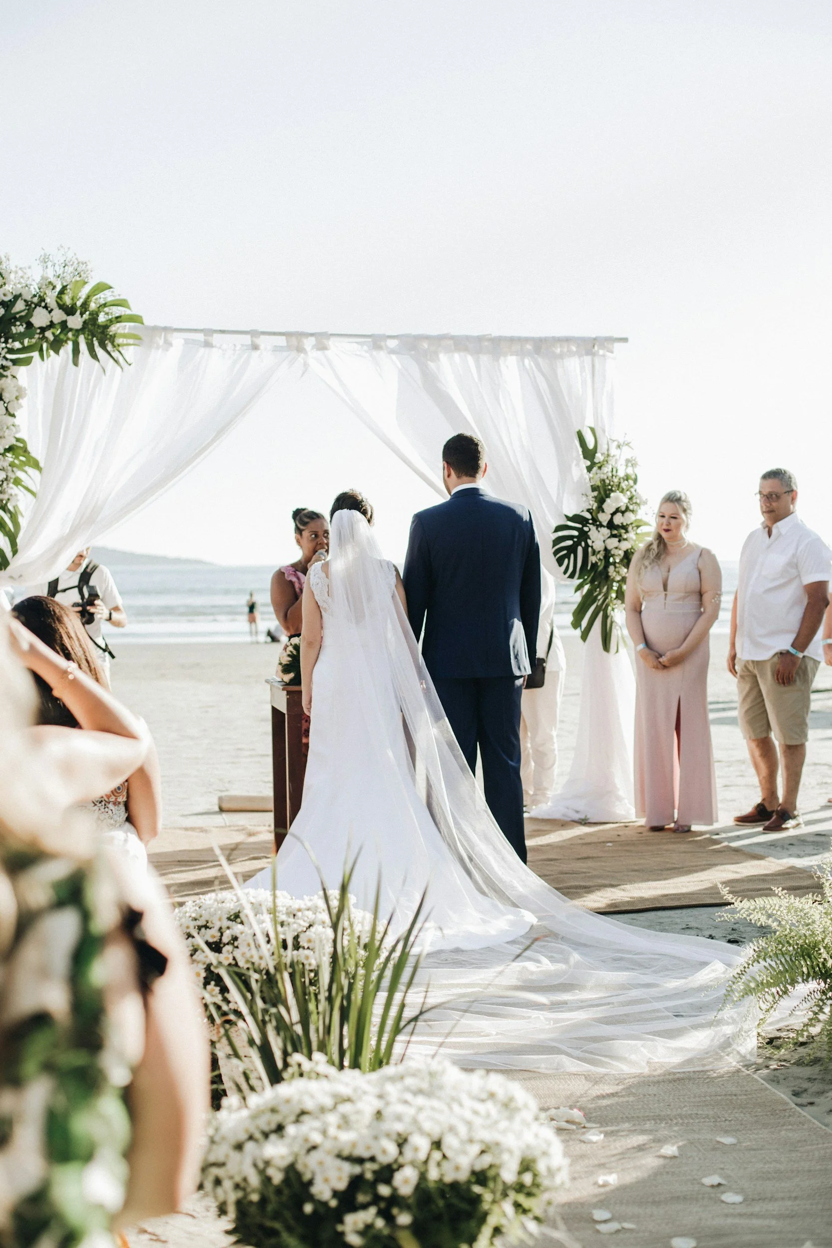 A beach wedding ceremony with a bride and groom standing under a white canopy adorned with flowers, facing each other, with guests and ocean in the background.