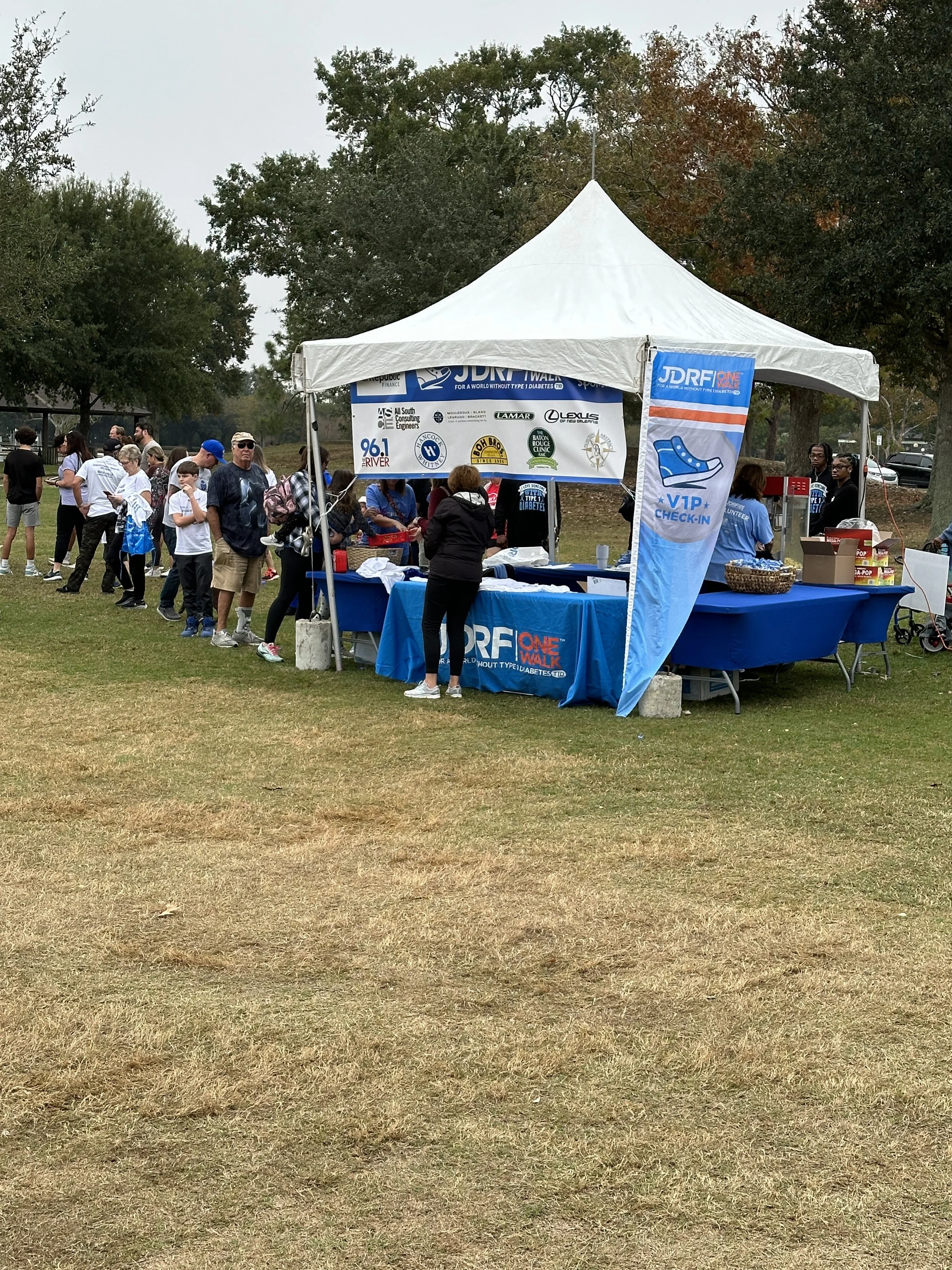 Outdoor event setup with a white tent, people standing around, and a grassy field.