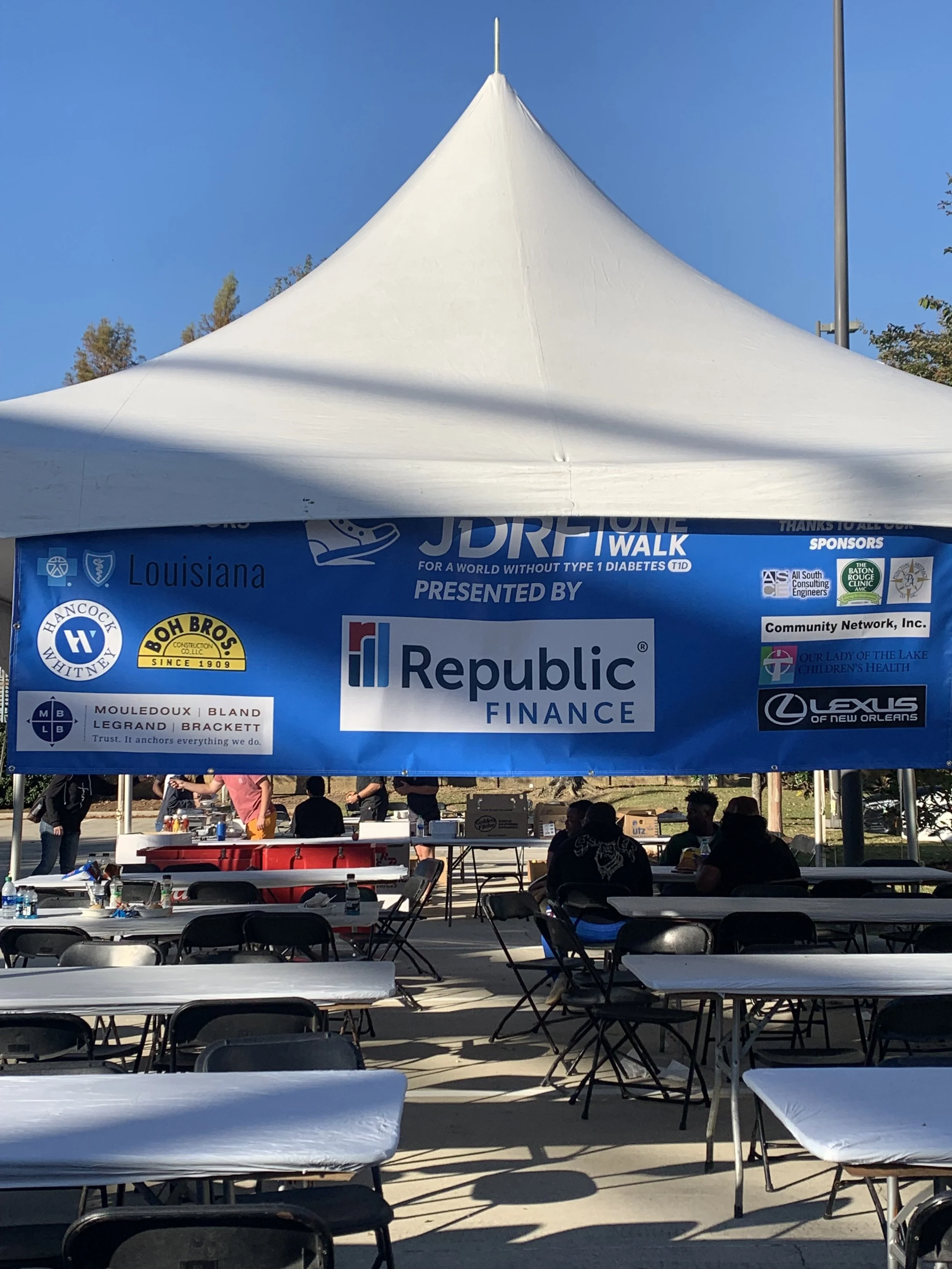 A large white event tent with a peaked roof, with a blue banner underneath displaying logos and text, set up in an outdoor area with tables and chairs, some people in the background, and trees and a clear blue sky overhead.