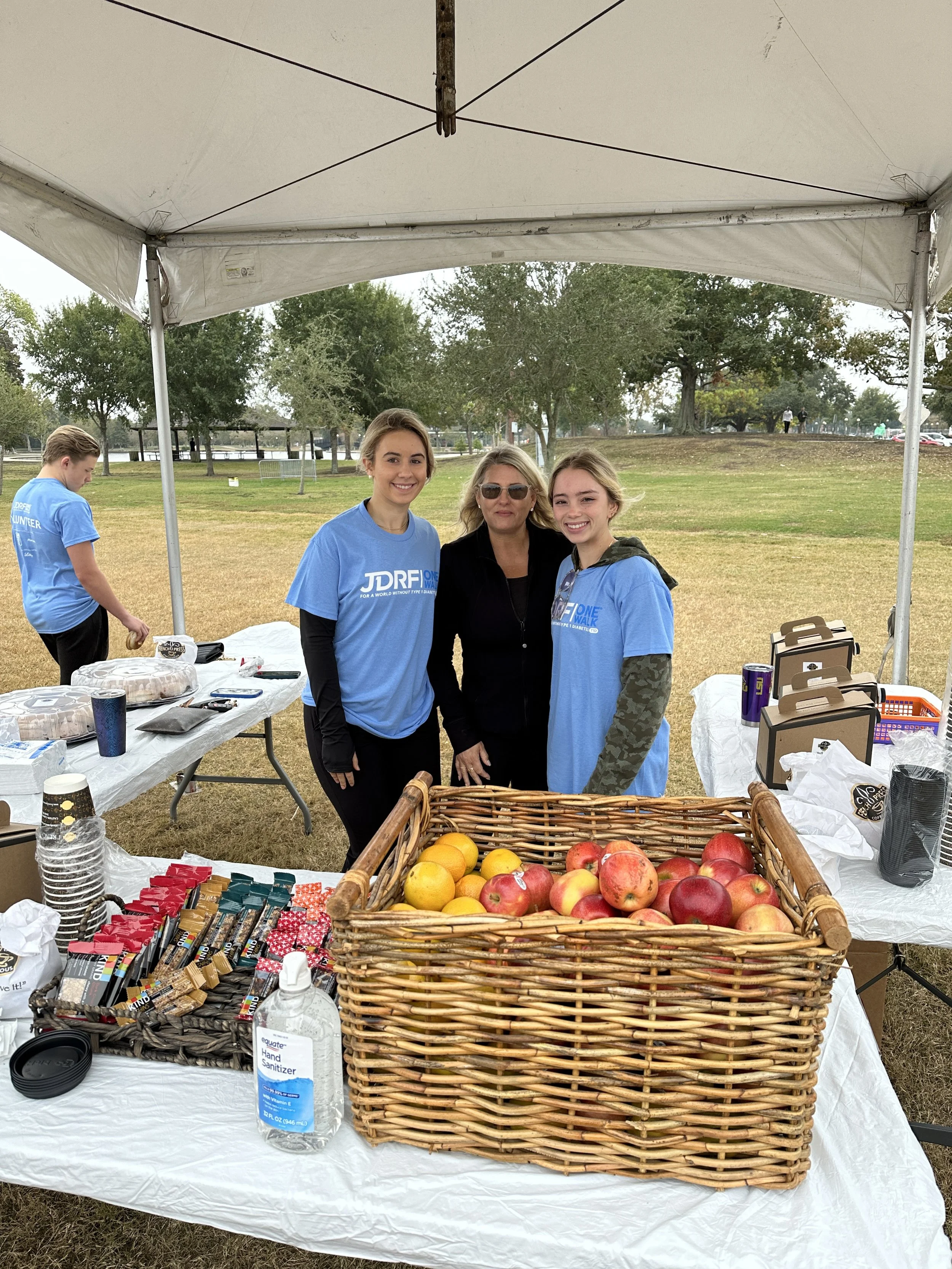 Four women under a white canopy at an outdoor event, with a table displaying apples, snacks, and hand sanitizer. One woman is centrally positioned, smiling, and three others are nearby. A park with grass, trees, and a few people walking in the backgr