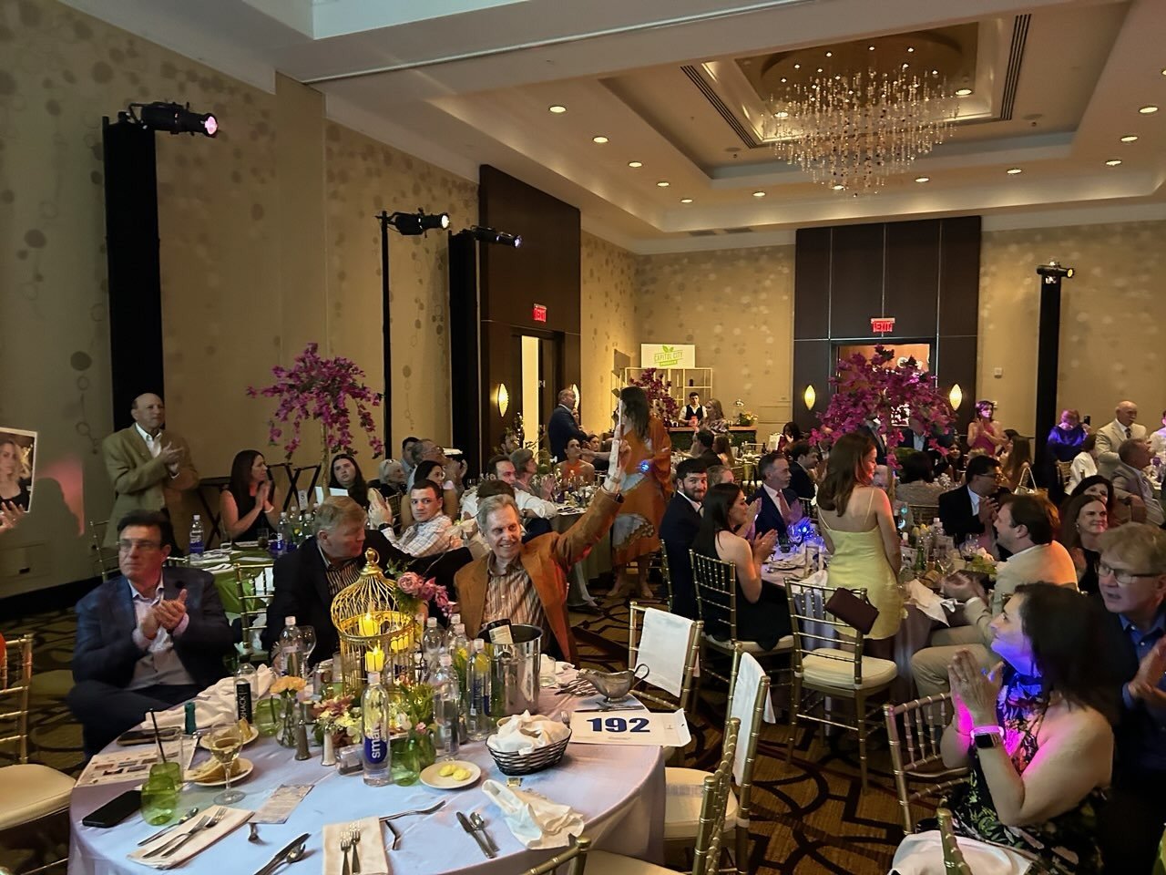 A large banquet hall filled with people celebrating, with decorated tables, floral centerpieces, and a chandelier, during a festive event.