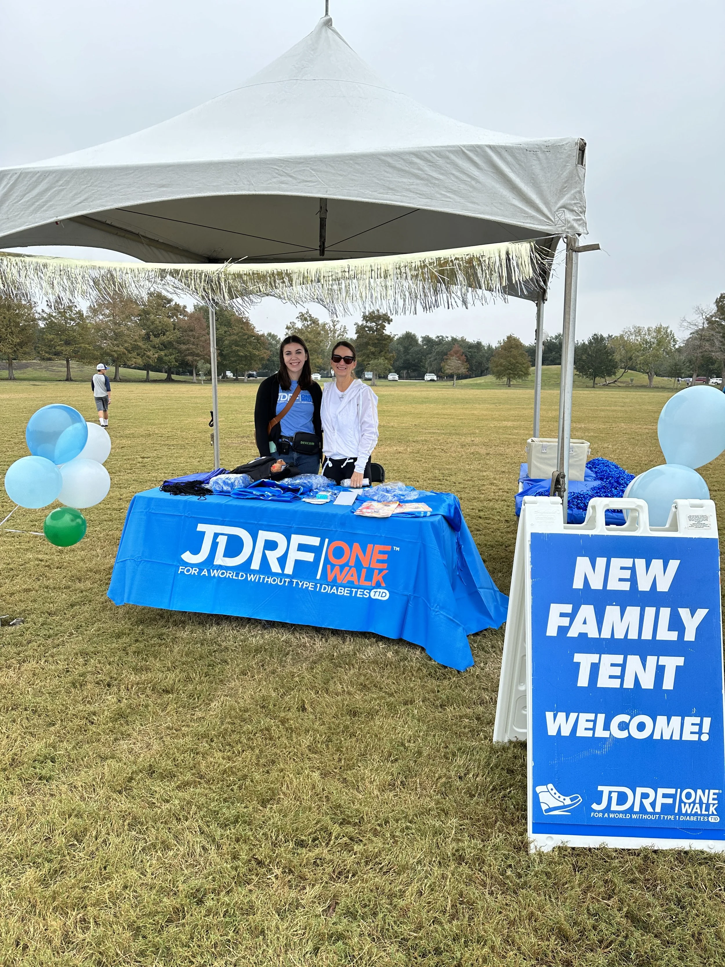 A booth set up under a white canopy tent at a park for JDRF One Walk, promoting awareness for Type 1 Diabetes. The table is covered with a blue cloth with the JDRF logo and information. There are balloons and a sign welcoming families.