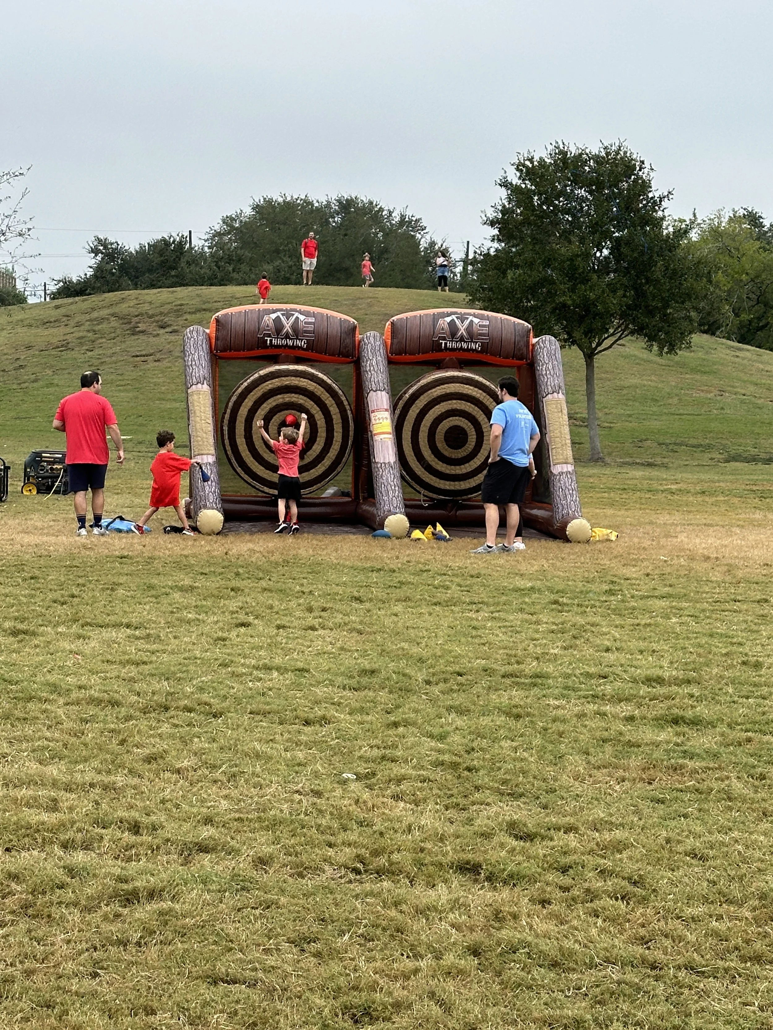 A group of children and adults playing a game of rollerball or target throwing at an outdoor park, with inflatable targets shaped like tree trunks and concentric circles, on a grassy field with a hill and trees in the background.