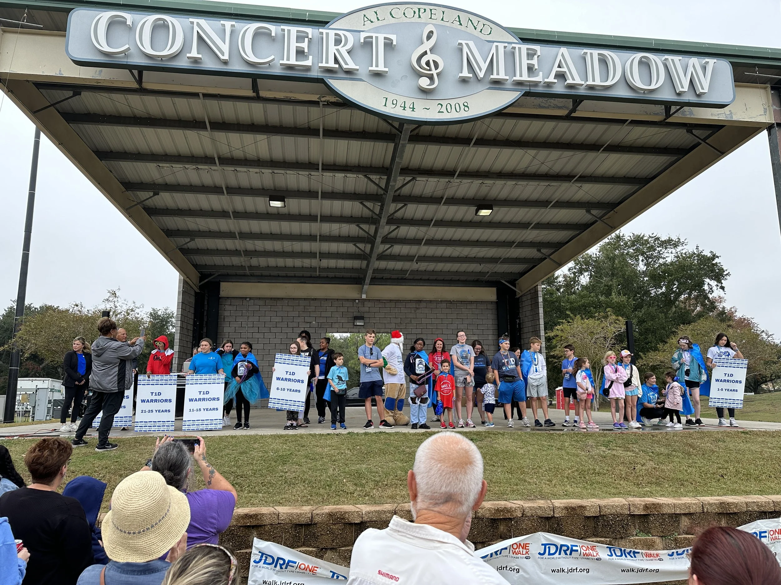 A group of children and adults on a stage at a concert and meadow event, with posters indicating different age groups and a large sign above reading 'Al Copeland Concert & Meadow 1944-2008', featuring people taking photos and watching.
