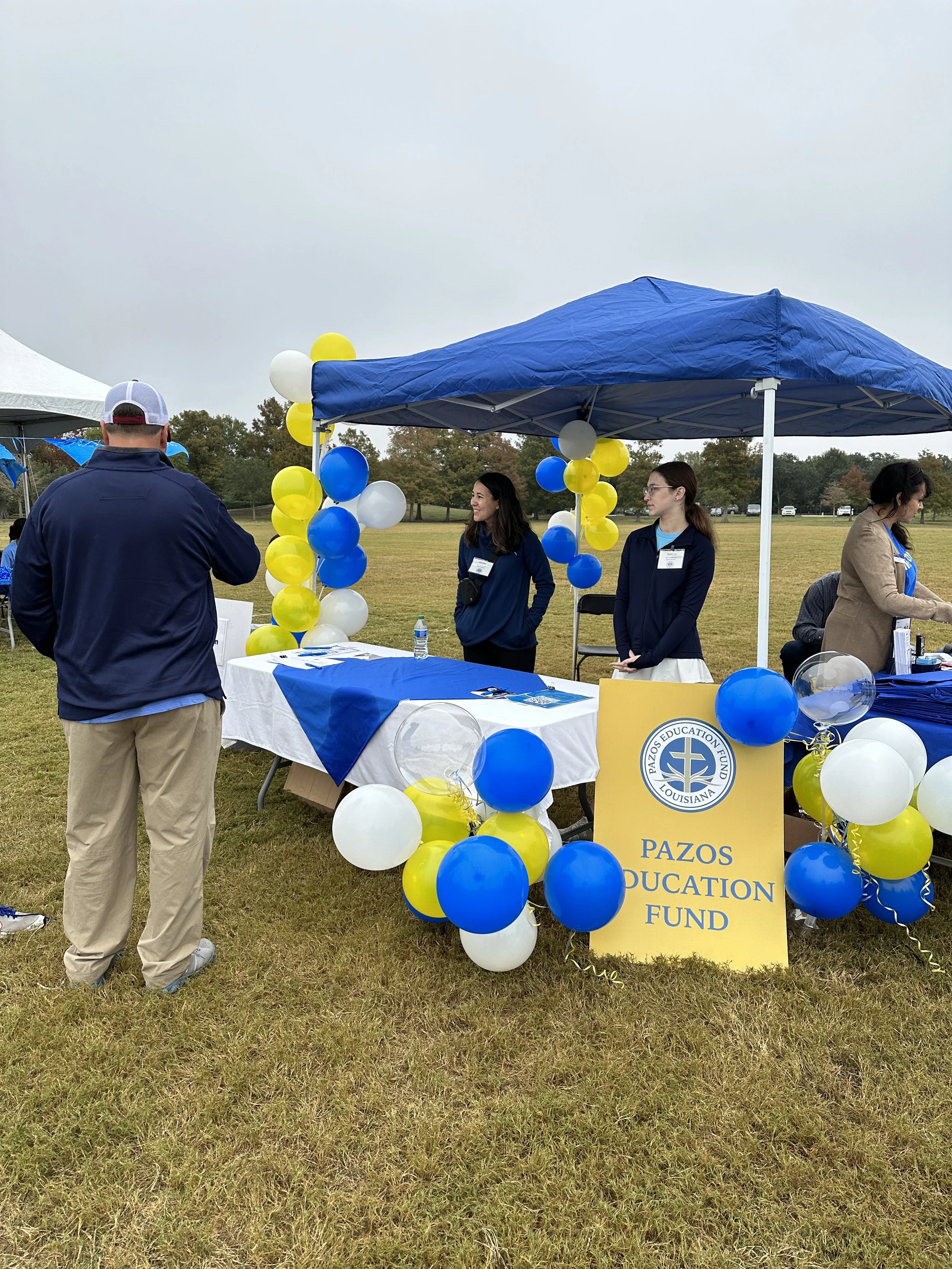 People manning booths at an outdoor event with yellow, white, and blue balloons, and a yellow sign for Pazos Education Fund on a grassy field with trees and a cloudy sky in the background.