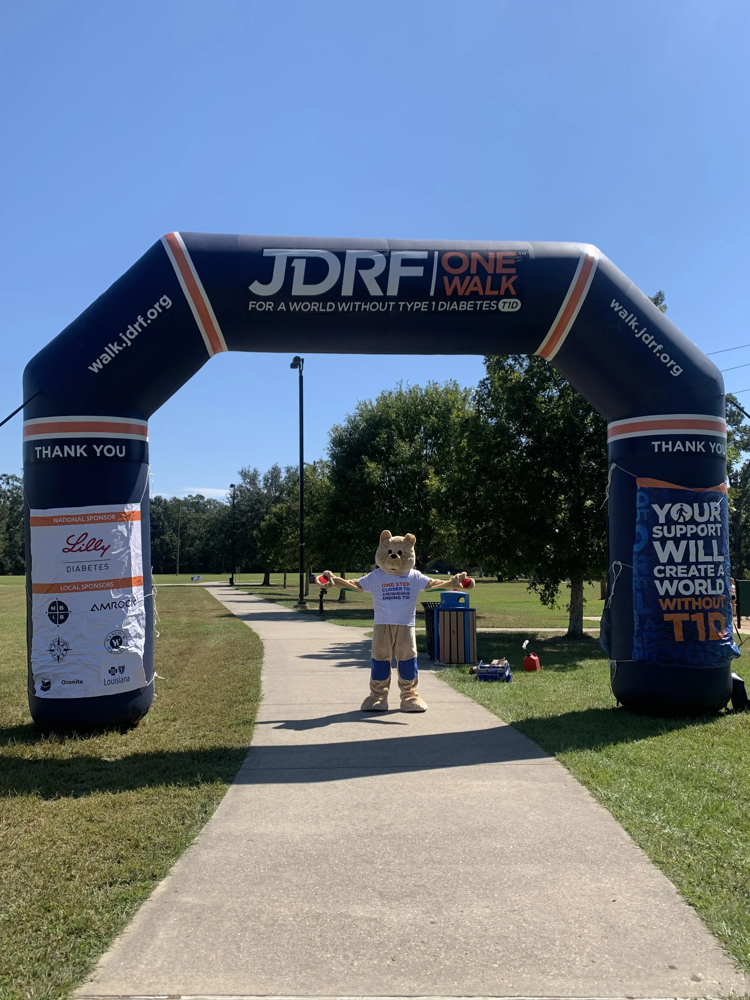 A person dressed in a bear costume standing at the finish line of a charity walk event called JDRF One Walk, with trees and a clear blue sky in the background.
