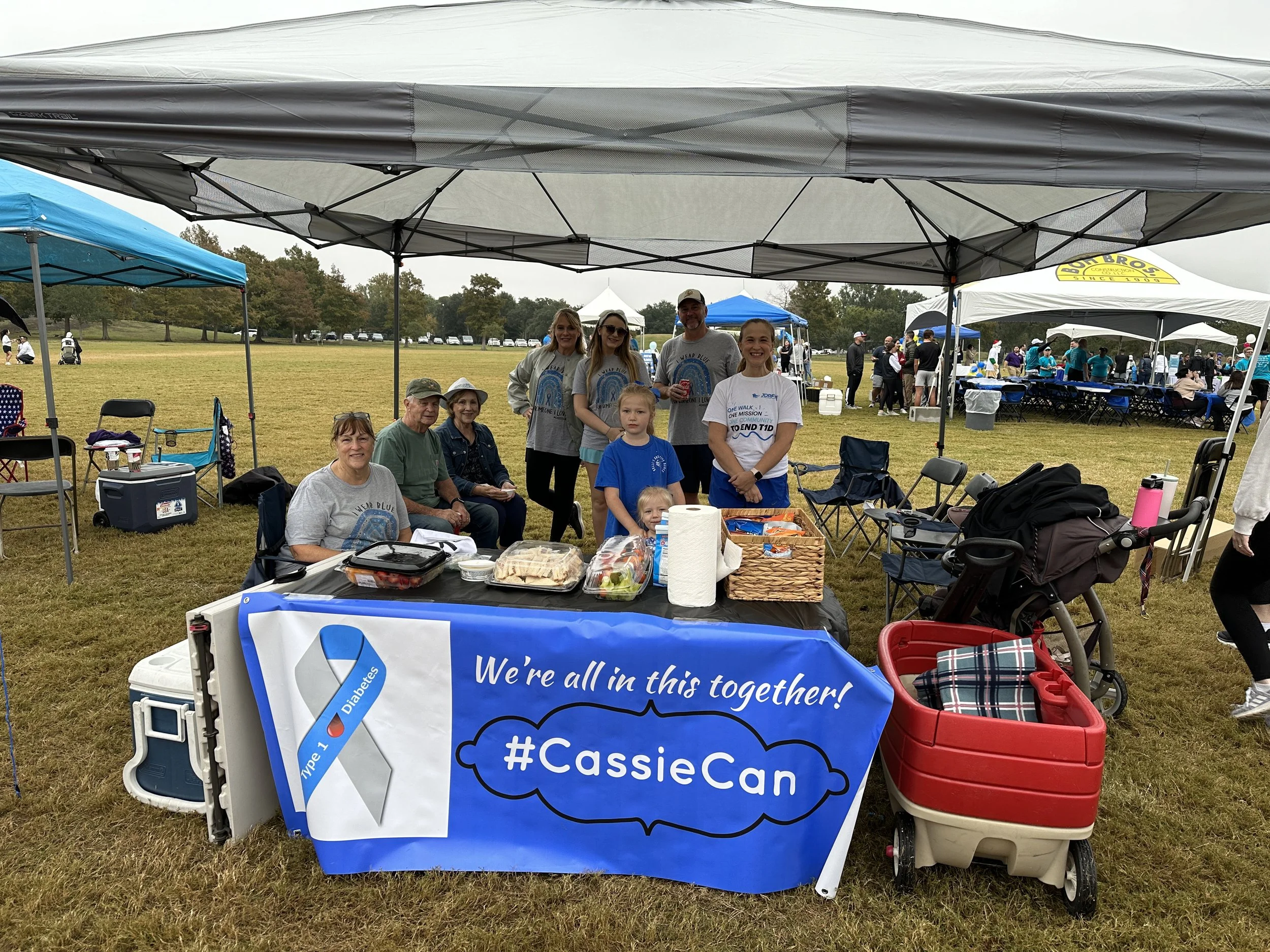 A group of people at an outdoor event under canopies, with a table containing food and drinks in the foreground, and a banner promoting awareness for diabetes with the hashtag #CassieCan.
