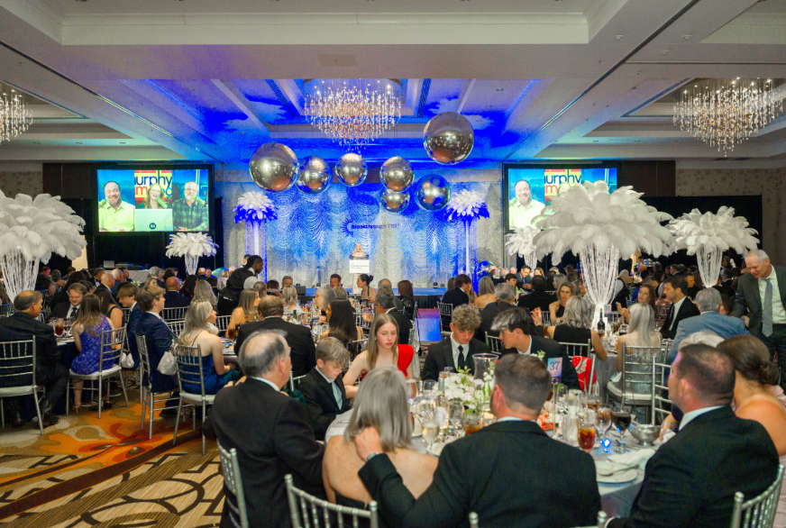 A large banquet hall decorated for a formal event with round tables, elegant centerpieces, and a stage with a speaker. The hall features elaborate lighting, chandeliers, and giant white floral arrangements. There are multiple screens displaying a presentation, and guests are dressed in formal attire, seated and listening to the speaker.