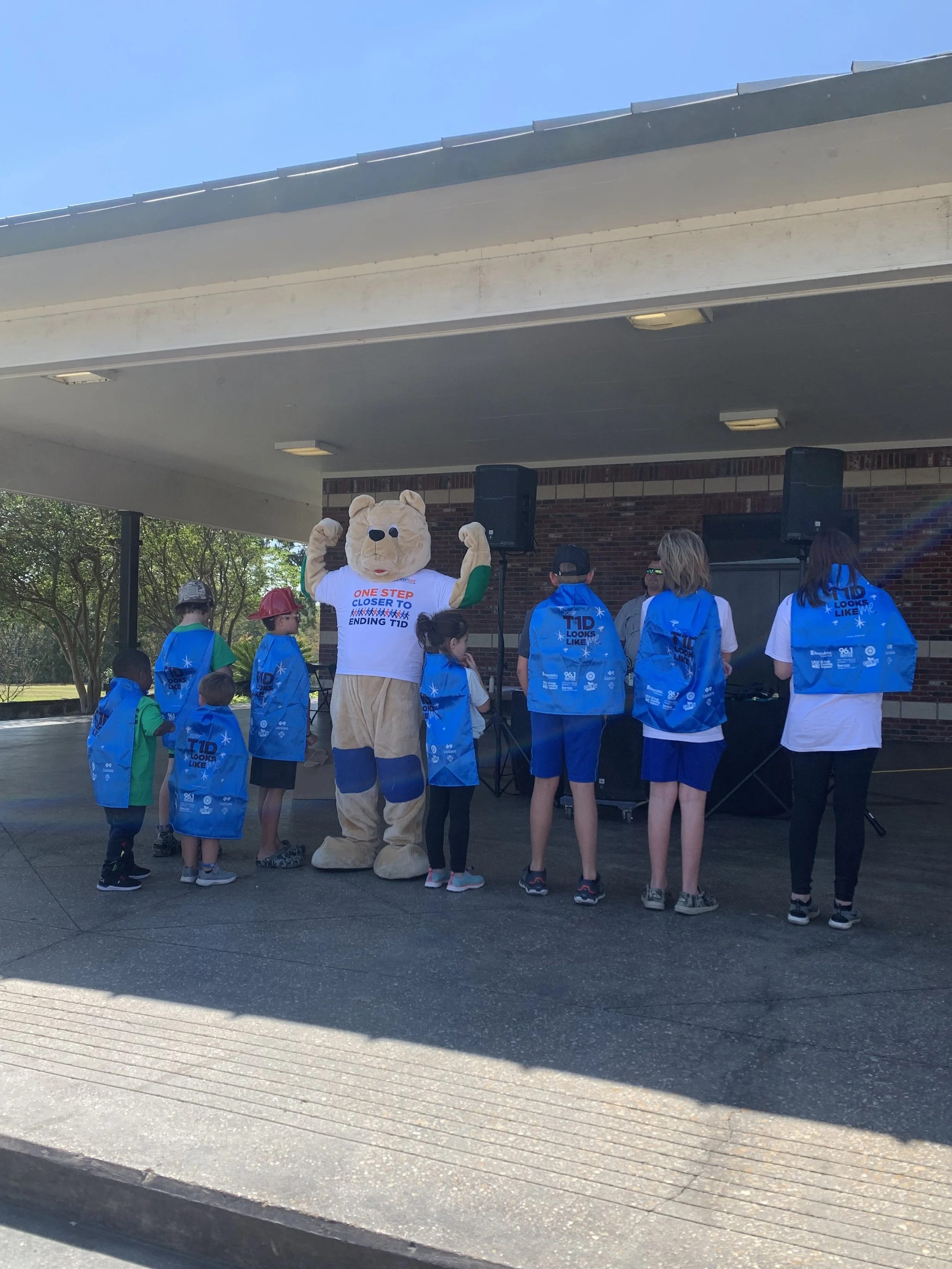 A group of children and adults gathered outdoors during daytime, with one person dressed in a bear mascot costume. The mascot is standing with arms raised, wearing a white shirt with text that mentions ending T1D. The children are wearing blue backpa