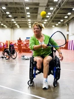 A woman in a wheelchair playing a court sport in an indoor sports facility, hitting a ball with a racket.