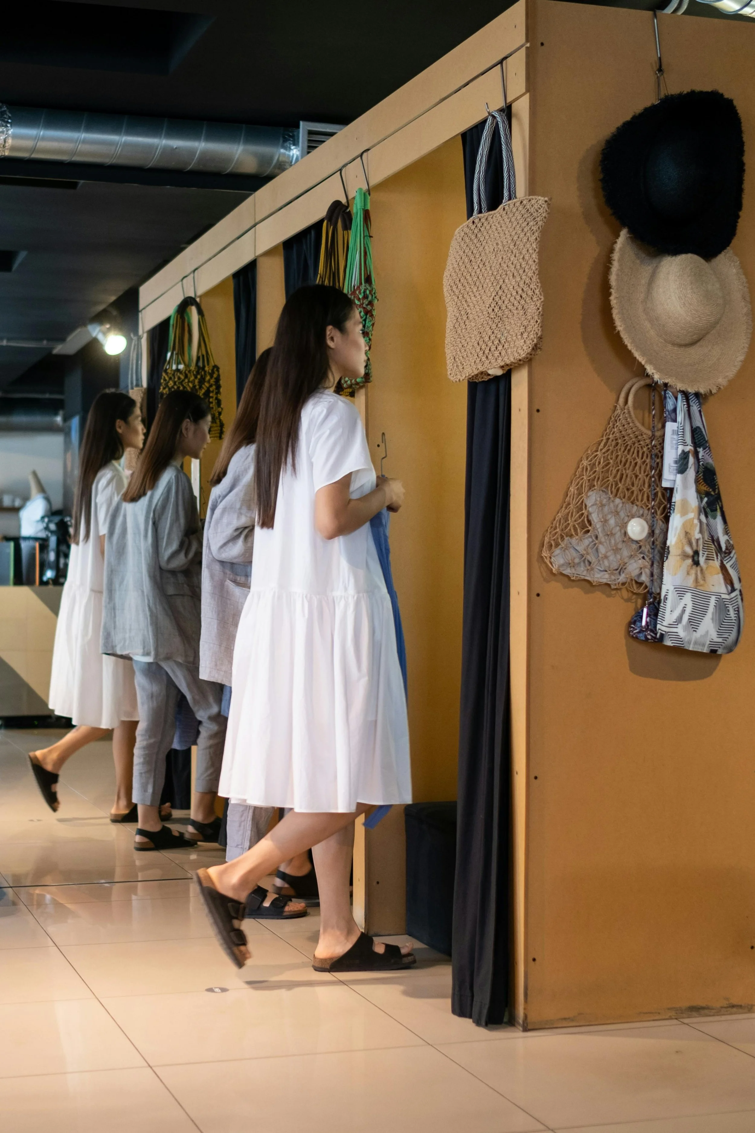 Multiple women in white dresses standing in line at a store, browsing straw hats and bags displayed on a wooden panel.