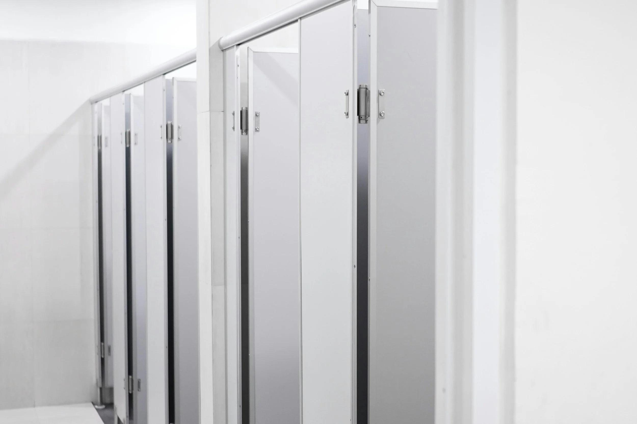 A row of white bathroom stall doors with metal handles and hinges in a public restroom.
