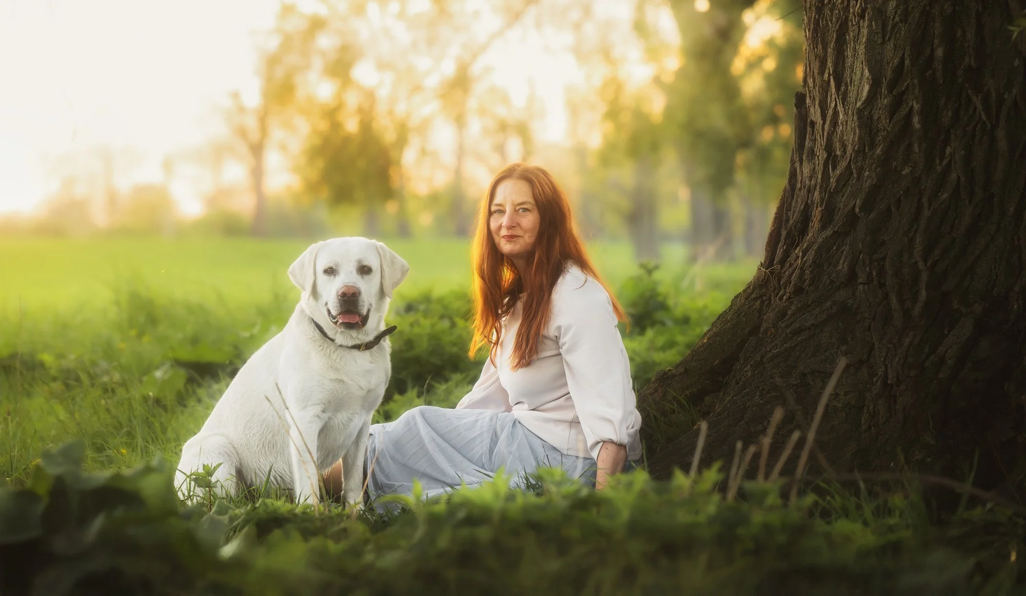 Eine Frau sitzt auf einer Wiese an einem Baum mit ihrem Hund und beide schauen in die Kamera. Im Hintergrund sieht man das goldene Licht des Sonnenuntergangs. März 2026.