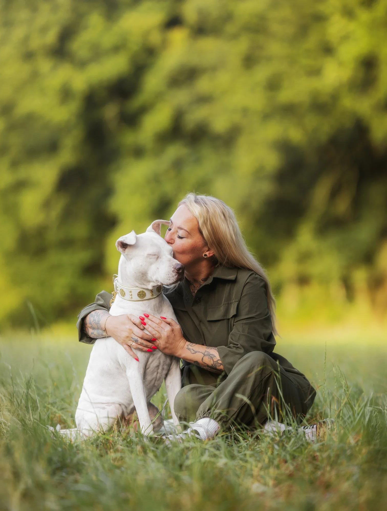 Eine Frau sitzt auf einer Wiese, umarmt ihren weißen Hund und gibt ihm einen Kuss. Der Hund hat dabei die Augen geschlossen und sieht aus, als würde er den Moment sehr genießen.