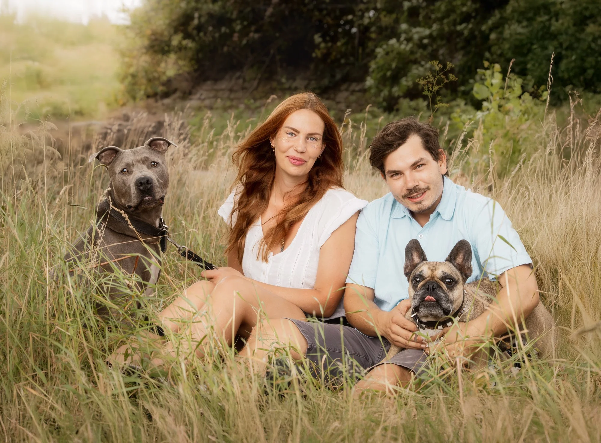 Ein Paar sitzt mit ihren beiden Hunden während eines Outdoor Fotoshootings im hohen Gras und alle schauen in die Kamera. Januar 2026.