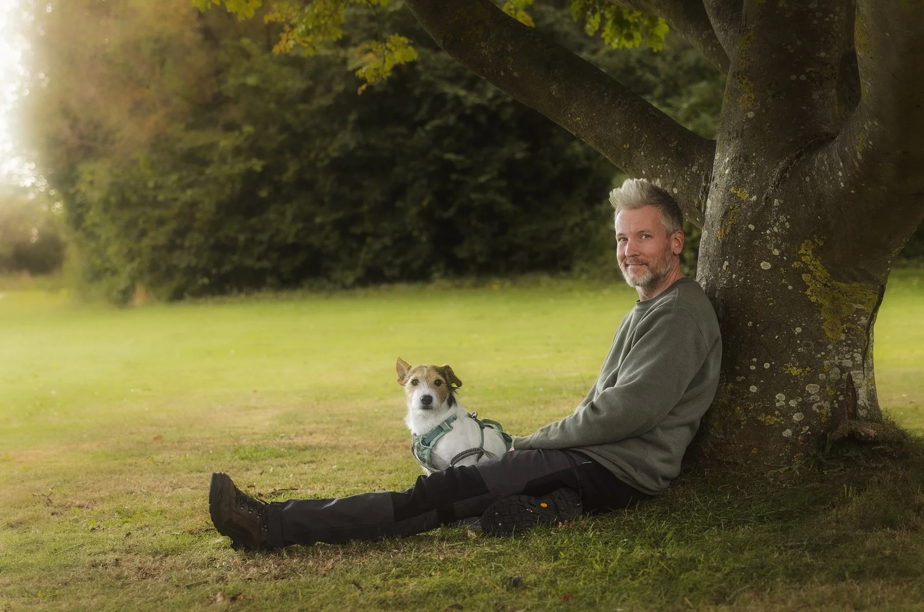 Ein Mann sitzt mit seinem Hund an einem Baum während eines Outdoor Fotoshootings für Hunde und ihre Menschen.