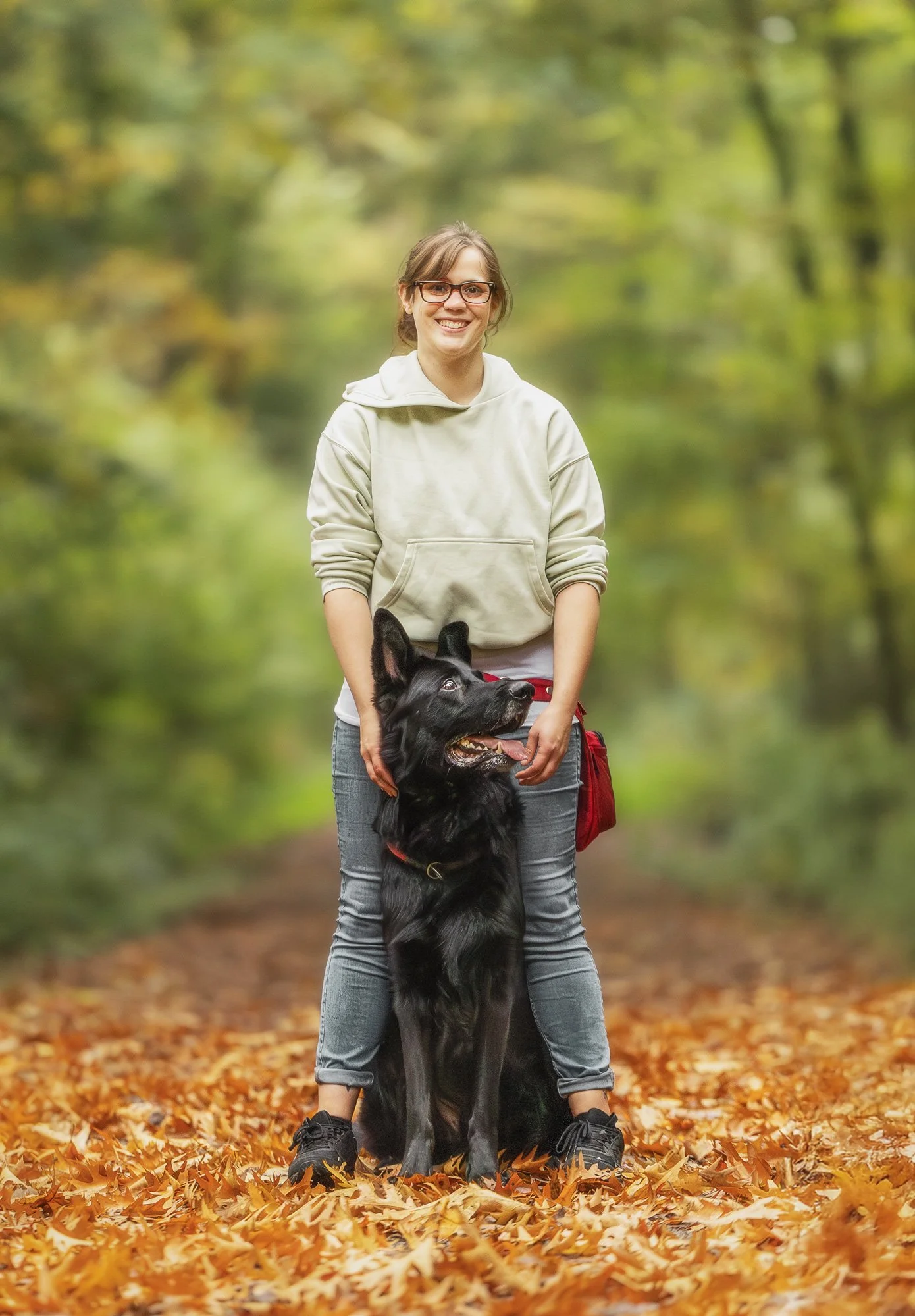 Eine Frau steht im herbstlichen Wald während eines Fotoshootings mit ihrem Hund. Ihr schwarzer Schäferhund steht dabei zwischen ihren Beinen und schaut in die Ferne.