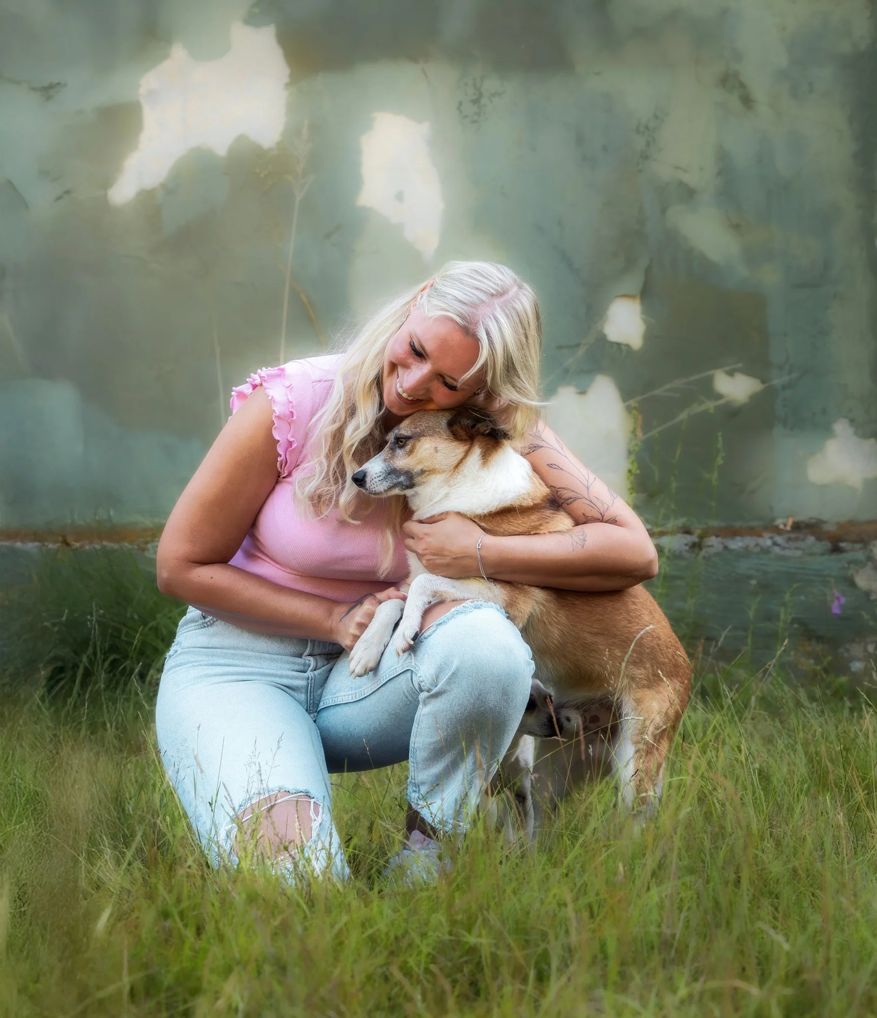 Eine Frau sitzt auf einer Wiese vor einem alten Gebäude und umarmt liebevoll ihren Hund.
