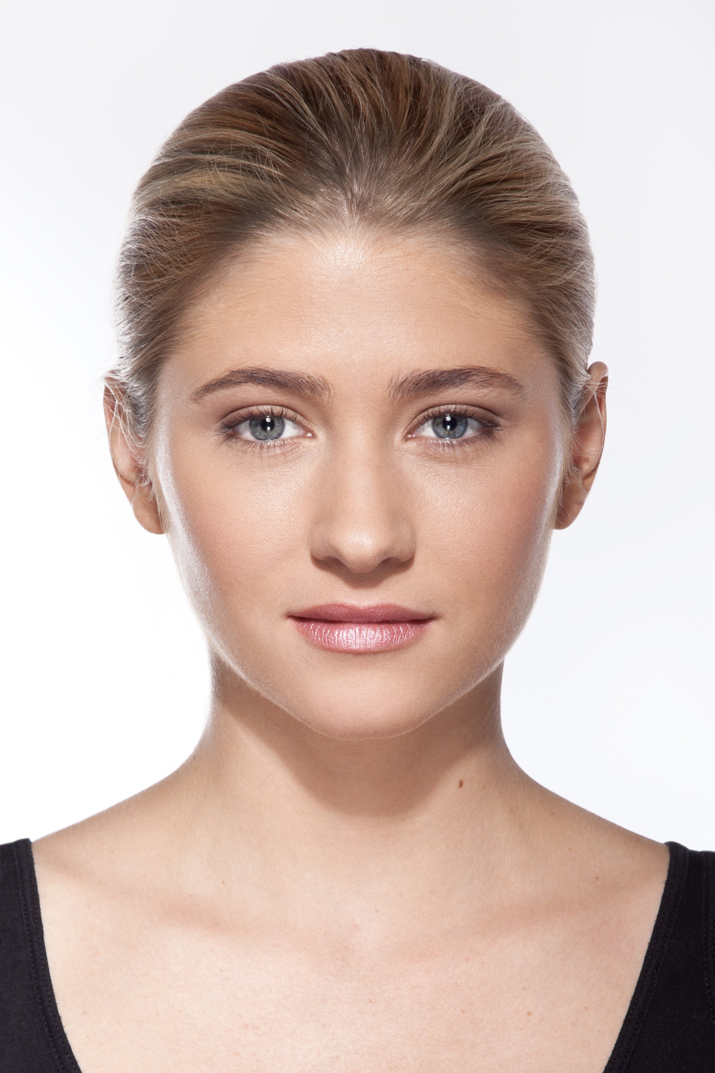 Close-up of a woman's face with clear skin, light makeup, and light brown hair pulled back, wearing a black top with a scoop neckline, against a plain white background.