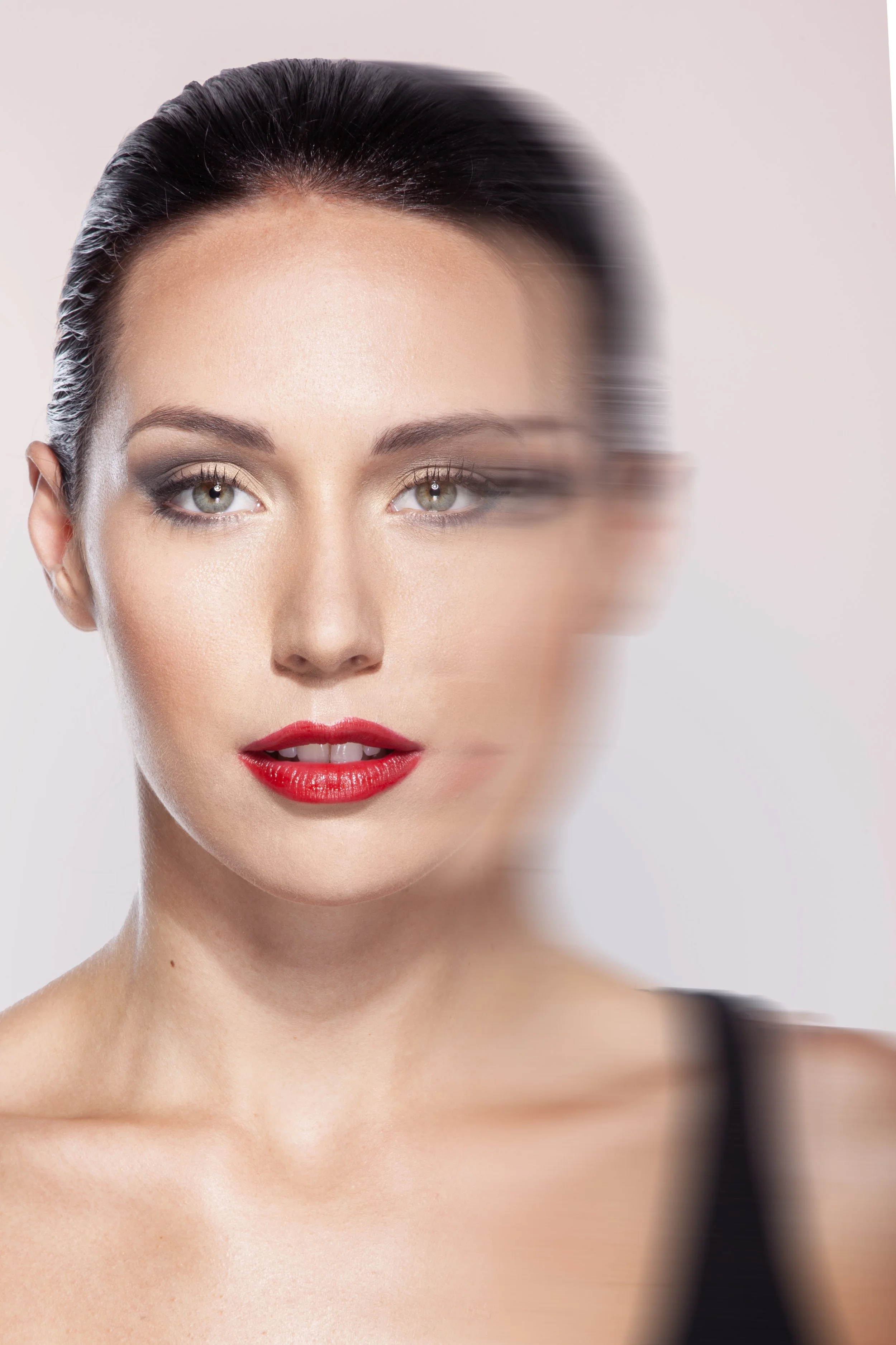 Close-up of a woman with fair skin, short dark hair, green eyes, red lipstick, makeup on her eyelids, and a neutral facial expression, with a slight motion blur.