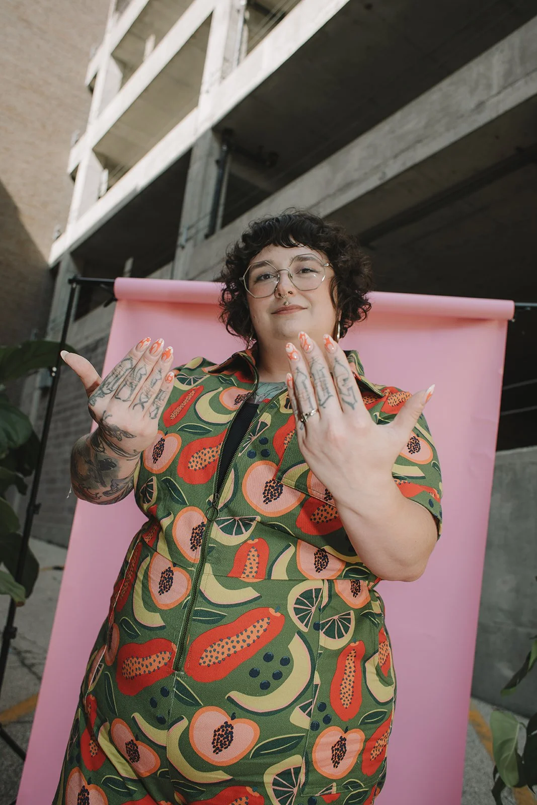 A woman with short curly hair and glasses shows her palms towards the camera, smiling, standing in front of a pink backdrop with a construction building in the background.