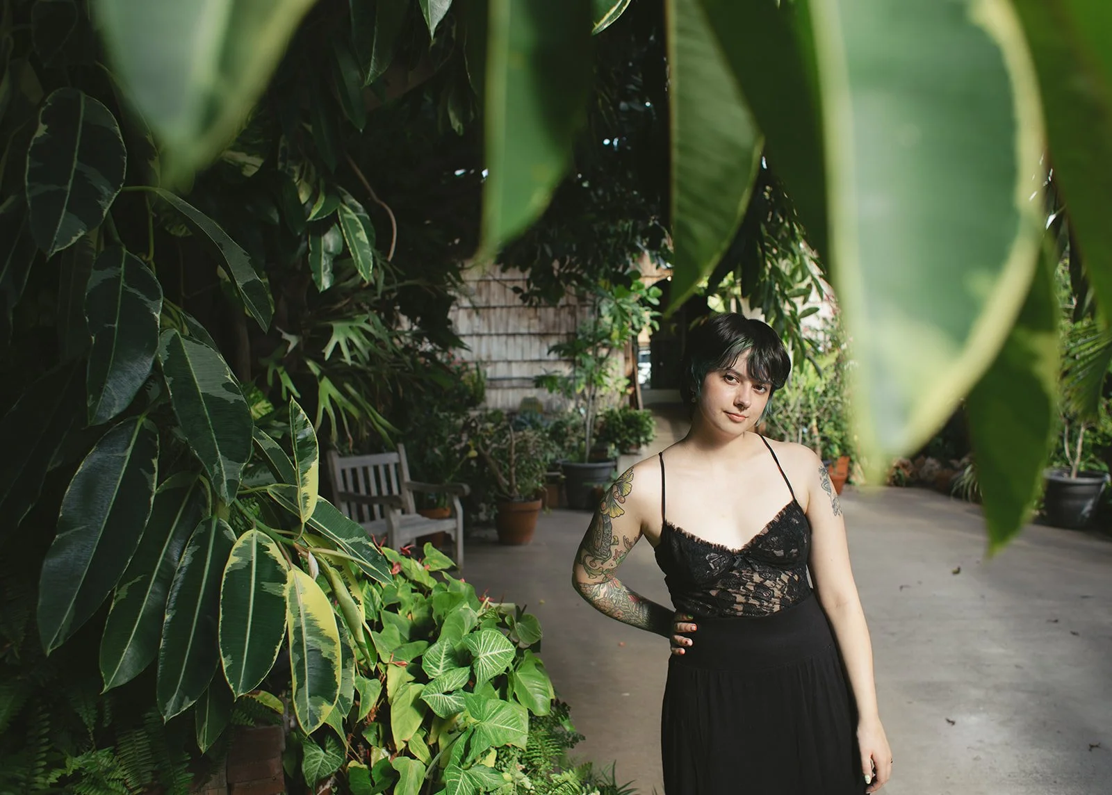 A young woman with tattoos on her arms and short black hair, wearing a black lace top and black skirt, standing in a lush, green garden with potted plants and a bench in the background.