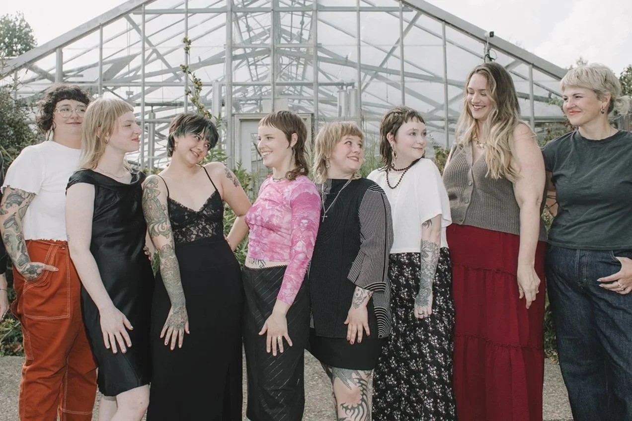 Group of women standing outside in front of a glass greenhouse, smiling and looking at each other.