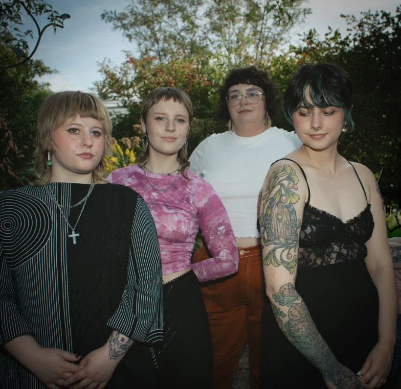 Group of six diverse women with tattoos and jewelry posing in front of a pink backdrop, some making finger heart gestures, holding flowers, and smiling.