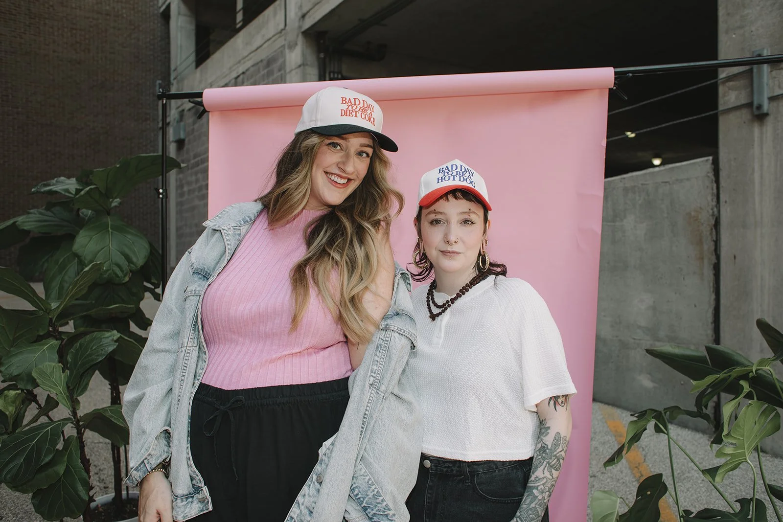 Two women with colorful hats smiling at a photo booth with a pink backdrop, surrounded by green plants.