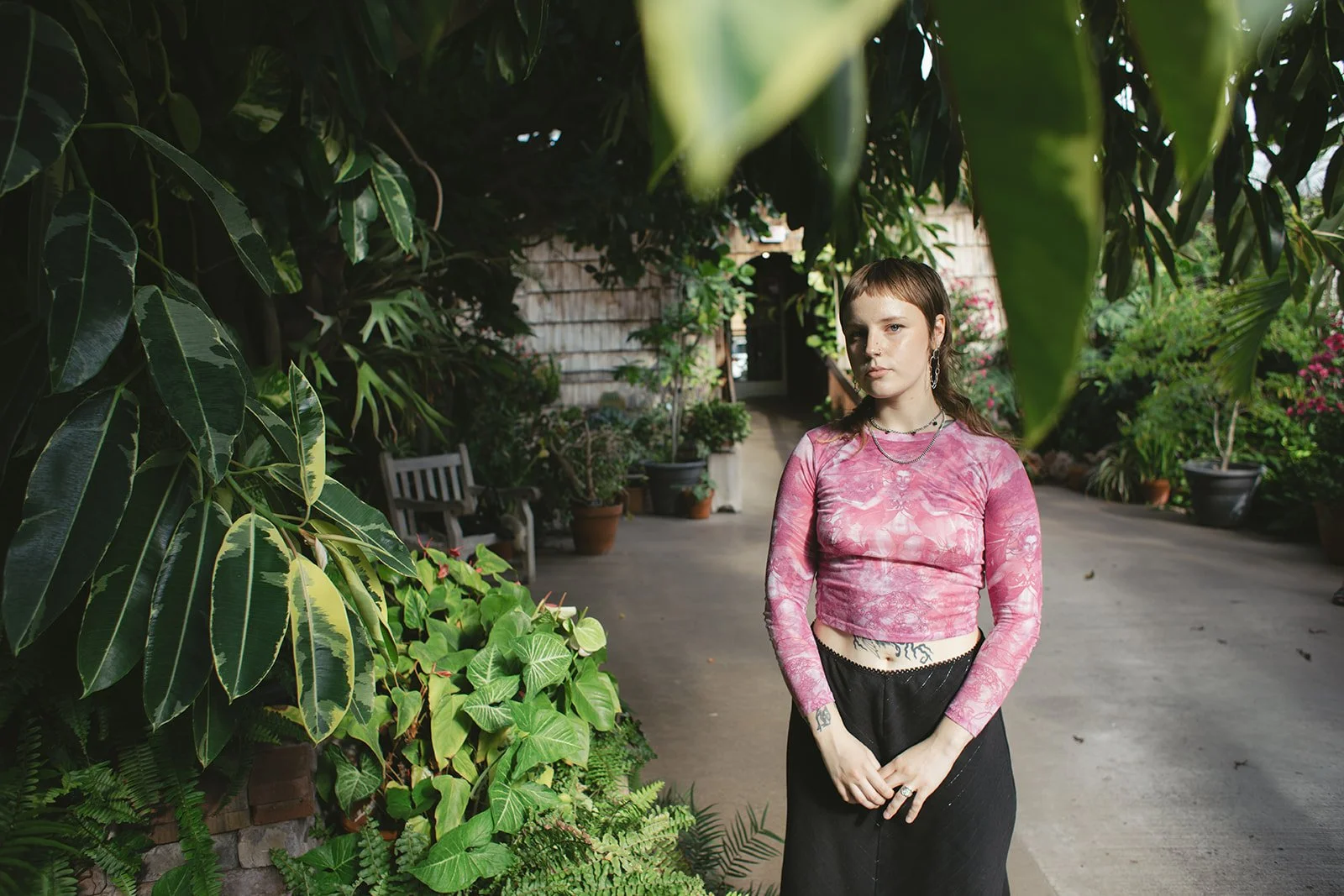 A young woman with short brown hair and tattoos, wearing a pink long-sleeve cropped shirt and black skirt, standing on a garden pathway surrounded by green plants and potted flowers.