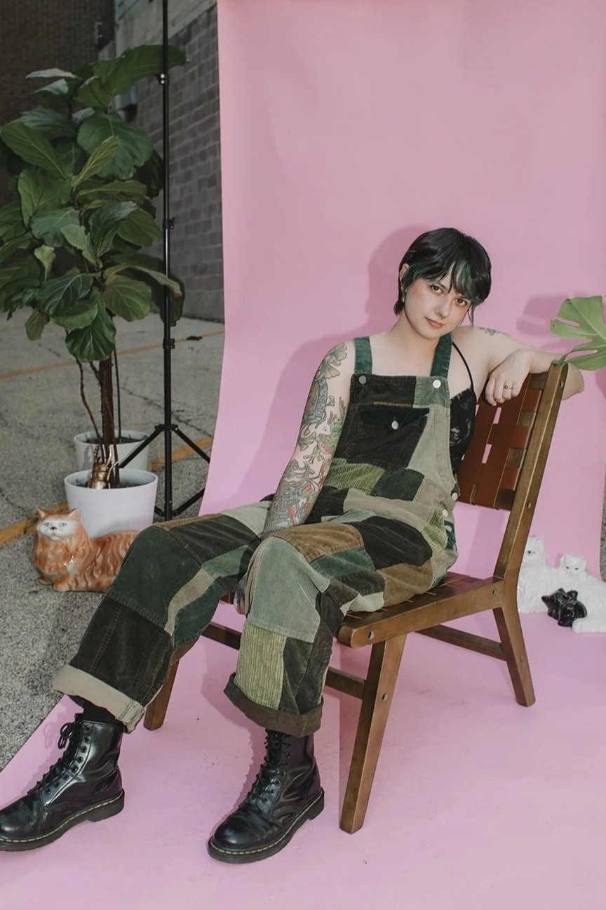 A young woman with short black hair and tattoos sitting on a wooden chair in front of a pink backdrop. She is wearing a patchwork denim jumpsuit with rolled-up pants and black combat boots. There are potted plants and cat figurines in the background.