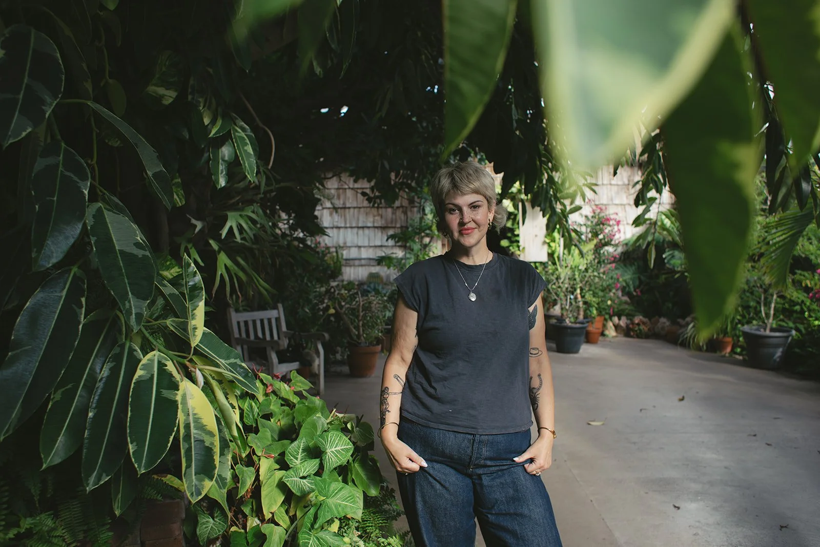 A woman standing outdoors in a garden surrounded by green plants and trees, wearing a dark t-shirt and jeans.