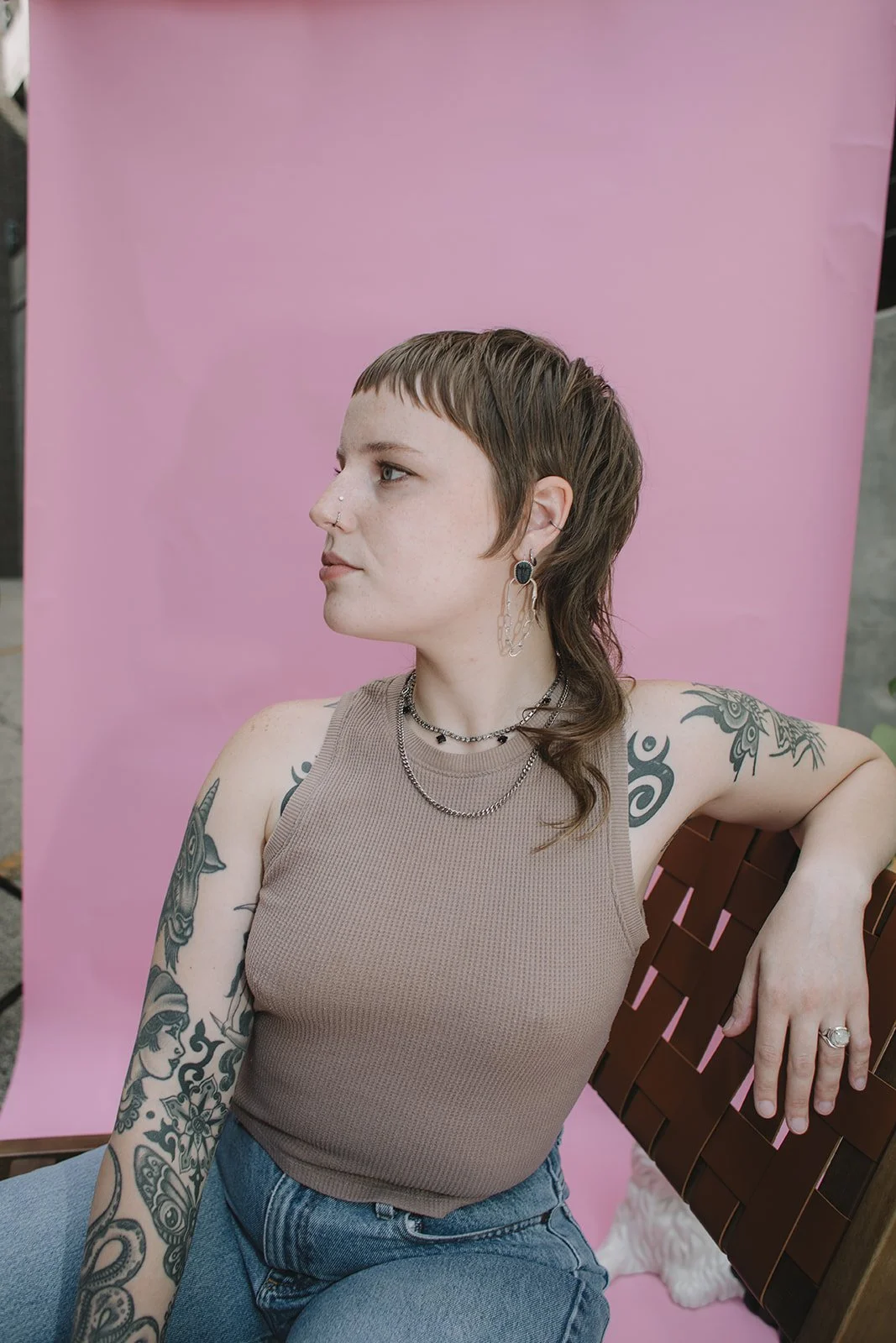 A young woman with short, layered brown hair and tattoos on her arms, wearing a beige tank top and blue jeans, sitting on a wooden bench against a pink backdrop.