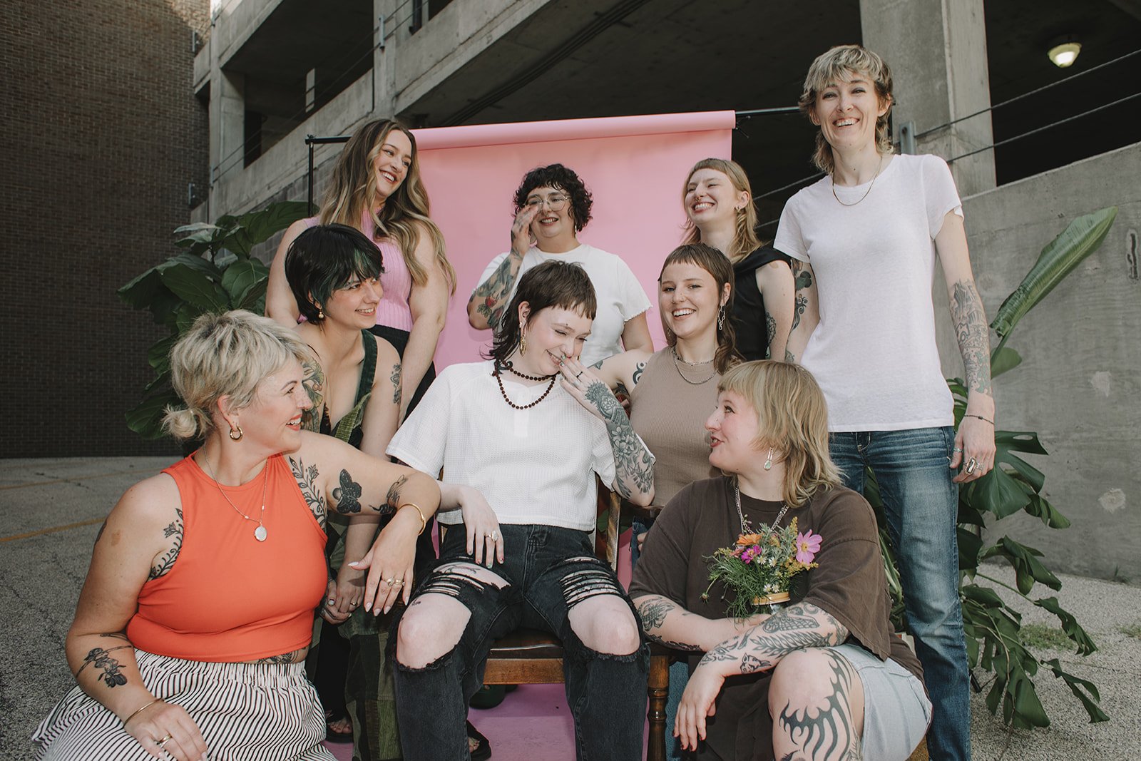 A group of eleven diverse women are gathered outdoors, smiling and enjoying each other's company. Some are seated, and some are standing, with a pink backdrop behind them and large green plants on either side.