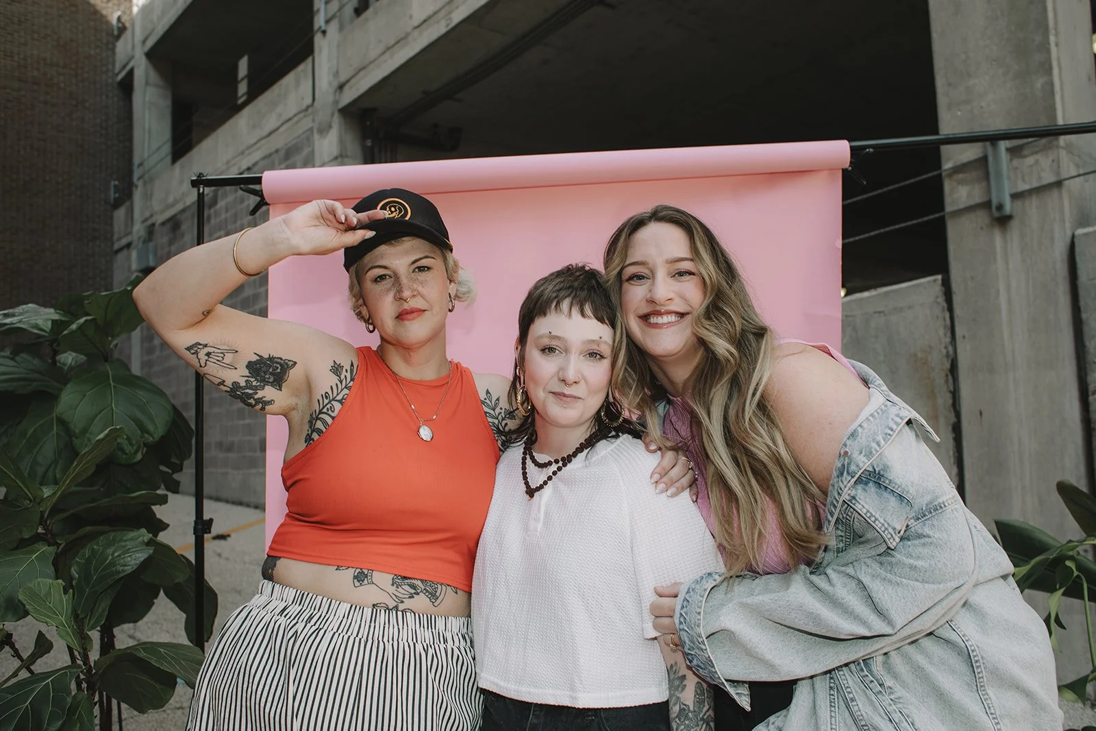 Three smiling women and one young person standing close together in front of a pink backdrop outdoors, with concrete buildings and green plants around them.