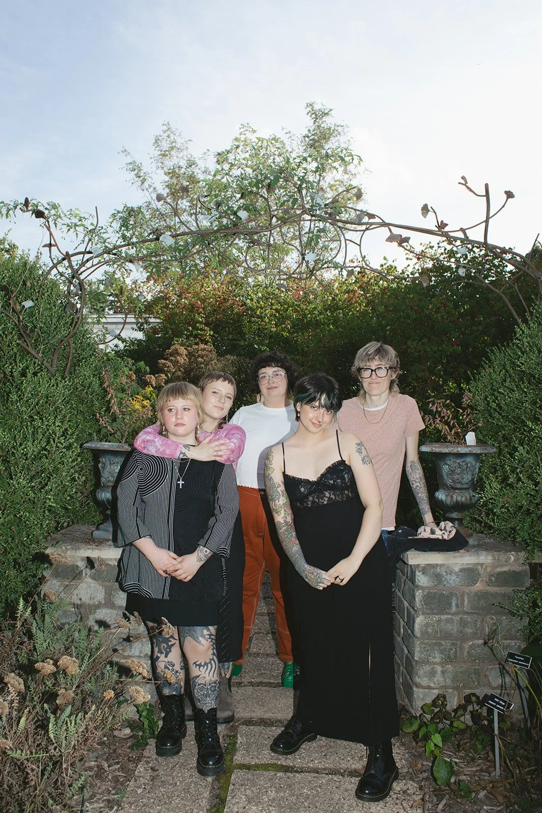 Five young women standing together outdoors in a garden, surrounded by greenery and stone planters, under a clear sky.