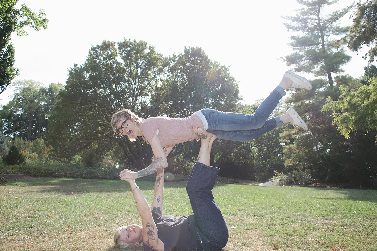 Two women doing acro yoga outdoors in a park on a sunny day, one lying on the ground and supporting the other who is balancing above her with arms extended downward.