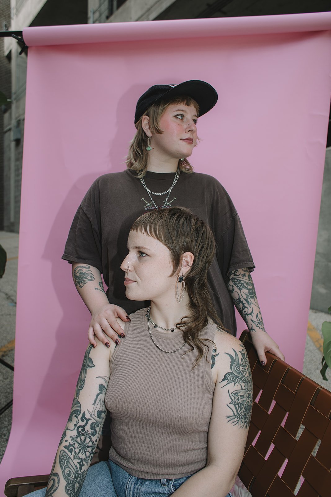 Two women with tattoos posing in front of a pink backdrop; one woman with short hair and a hat stands behind the seated woman with longer hair, both looking sideways.