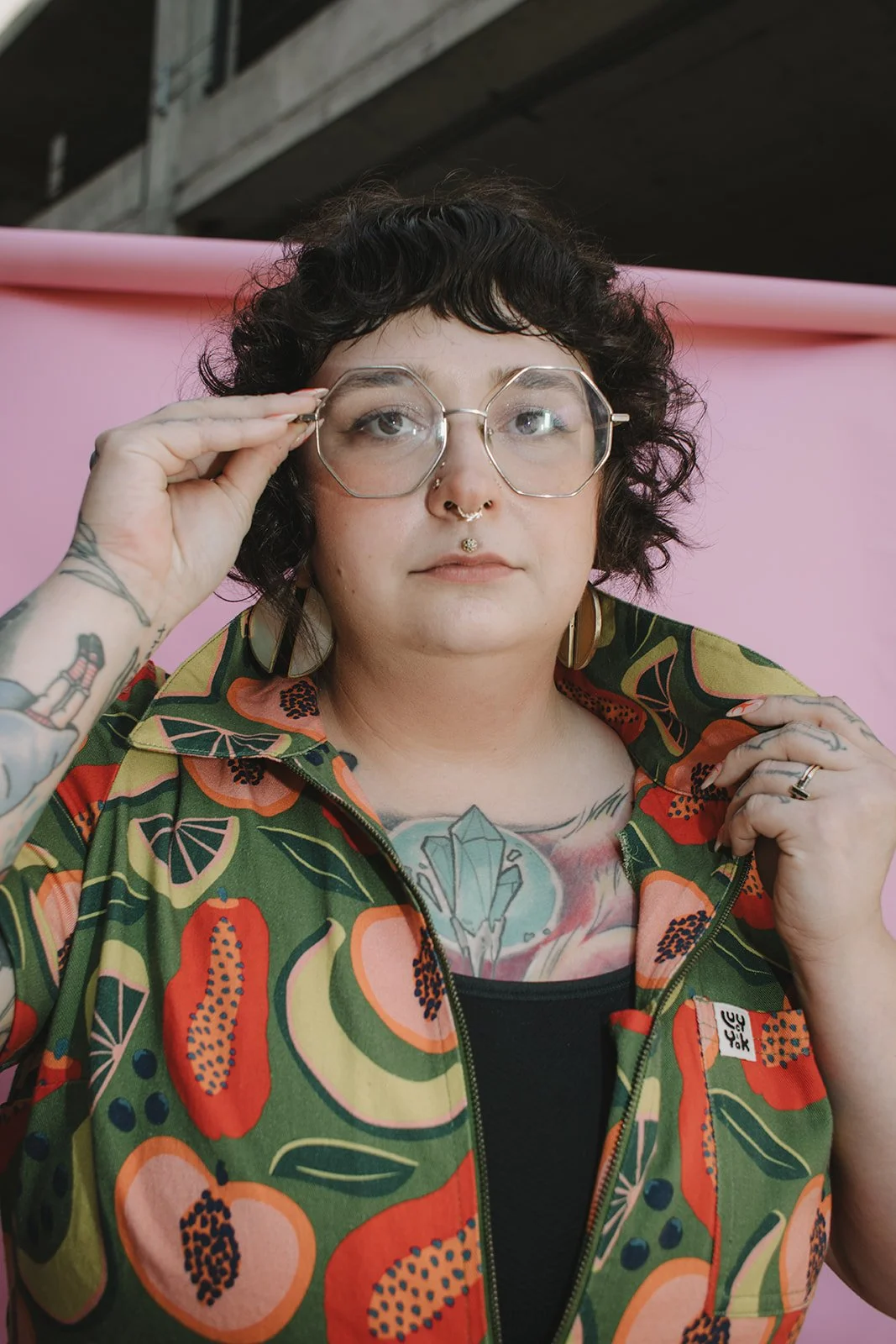 Portrait of a woman with curly hair, glasses, tattoos, and jewelry, wearing a colorful floral jacket, against a pink background.