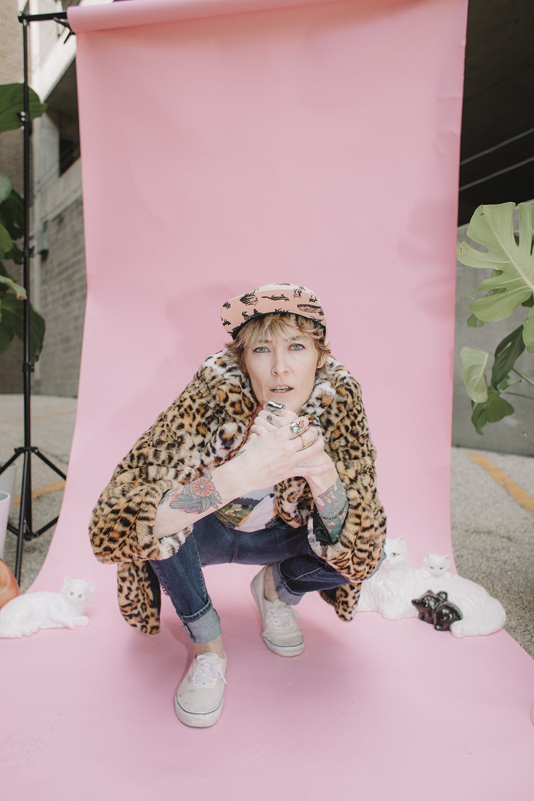 A woman with tattoos on her arms, blonde hair, and blue eyes squatting in front of a pink backdrop, wearing a leopard print coat, a patterned beret, and white sneakers, surrounded by white and black cat figurines.